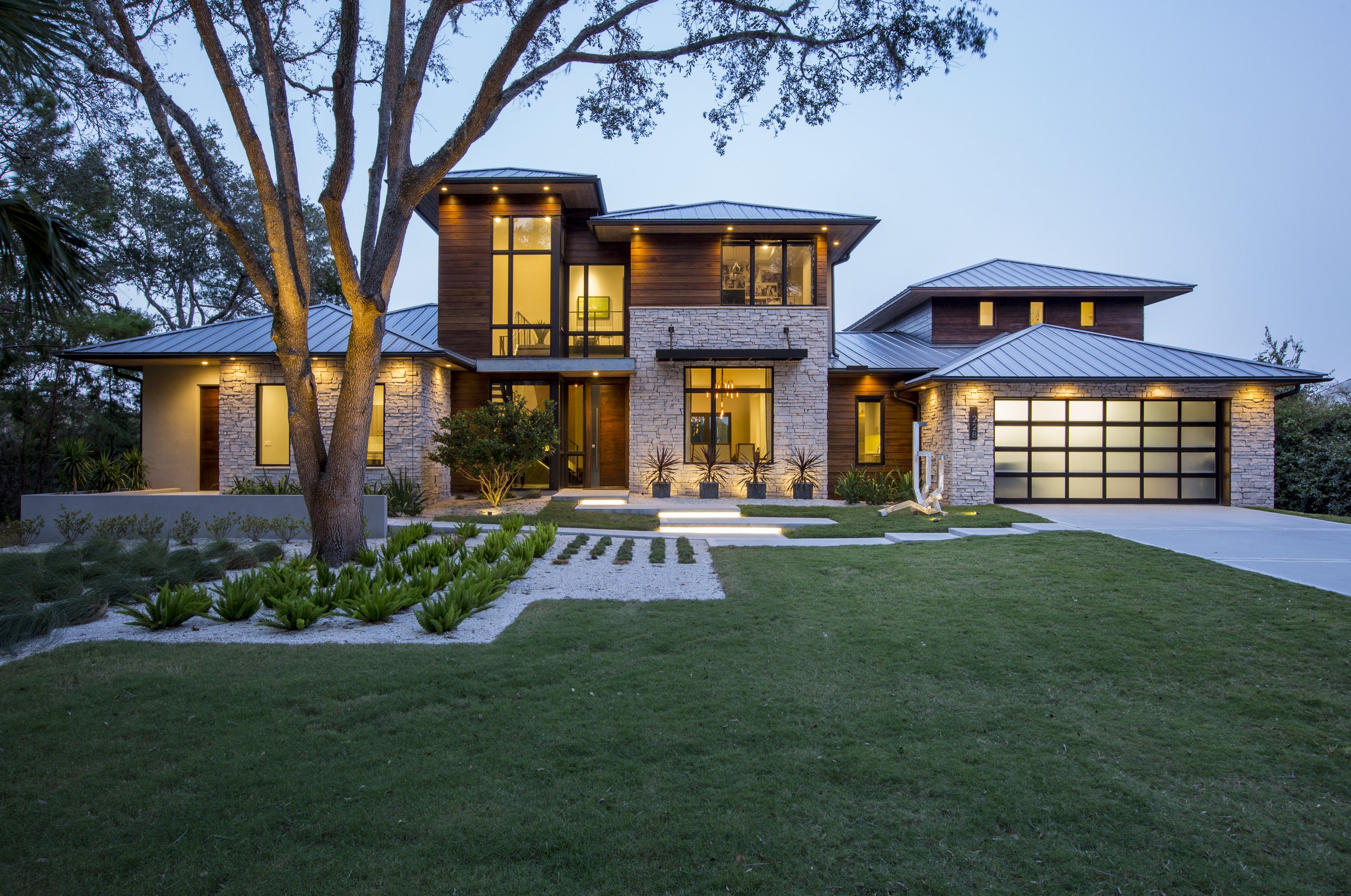 Modern two-story house with stone and wood exterior, large windows, and a metal roof; front yard with green lawn, small plants, and a pathway, surrounded by trees and lit up with exterior lighting at dusk.