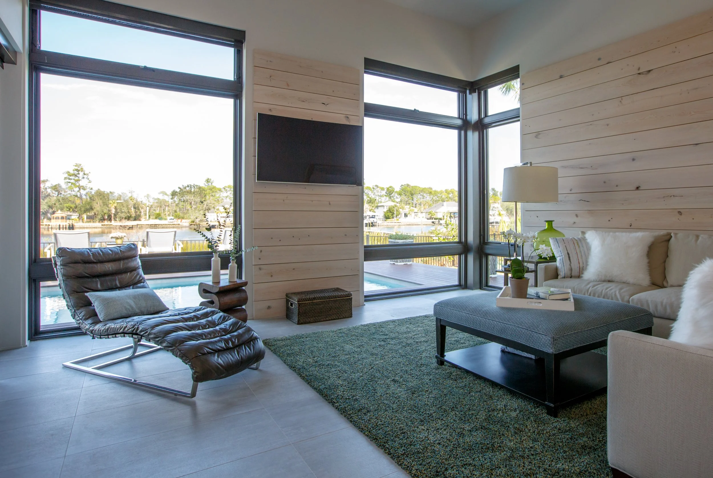 Modern living room with floor-to-ceiling windows overlooking water and trees. Contains a leather lounge chair, a sofa with pillows, a coffee table, a green glass vase, a lamp, and a TV mounted on a wood-paneled wall.