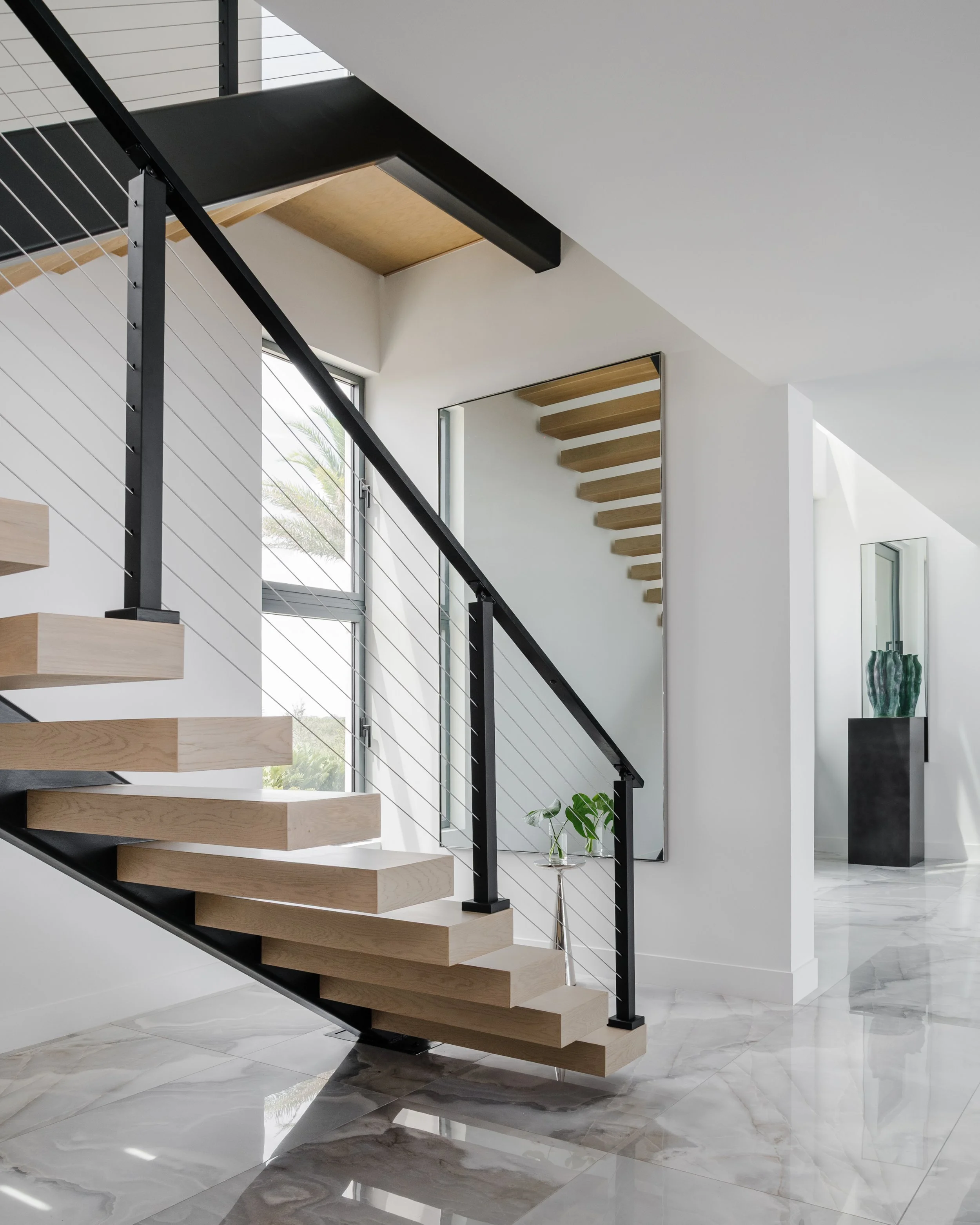 Modern staircase with wooden steps and black metal railing inside a bright home with white walls and marble flooring. Large windows let in natural light.