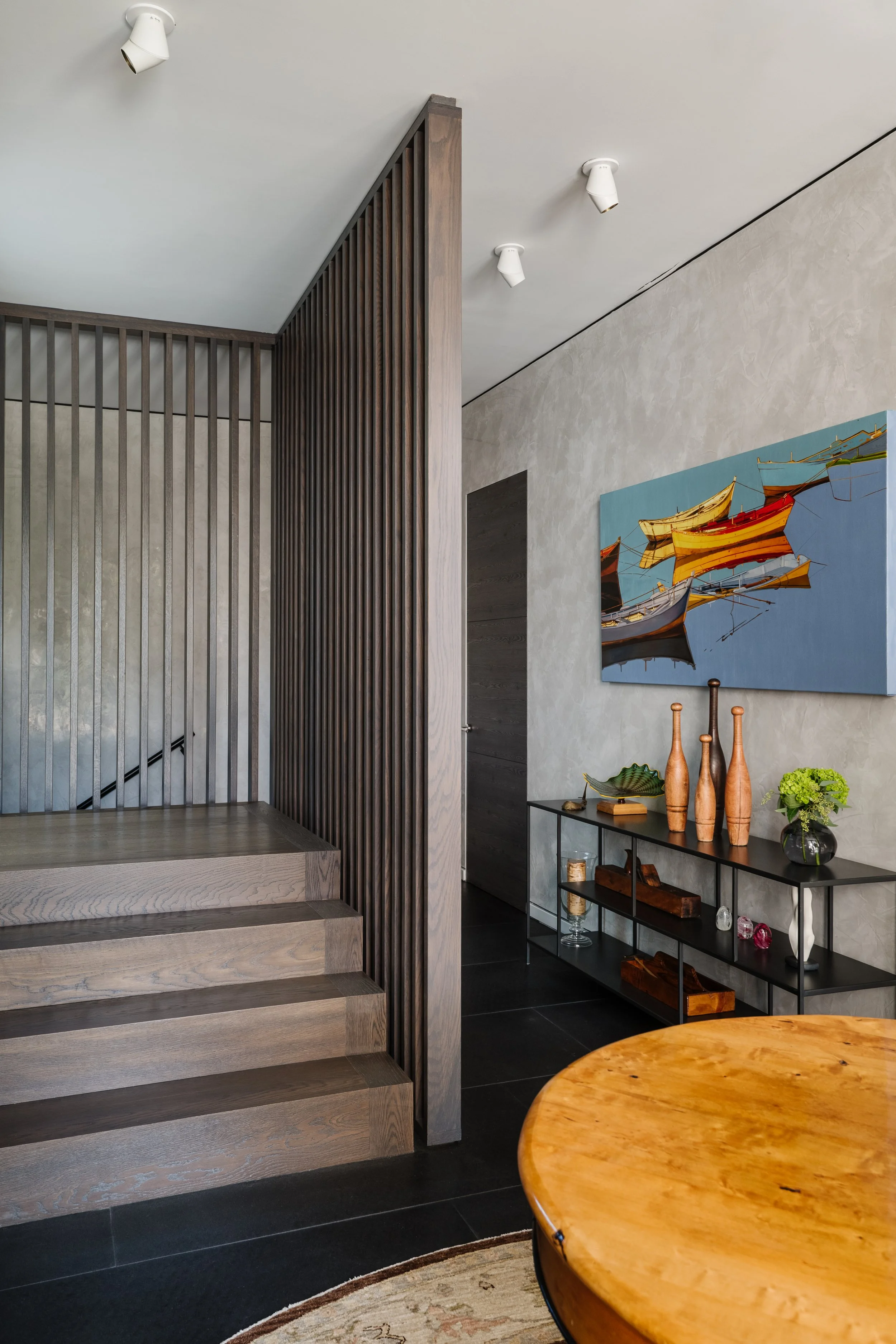 Interior of a modern home with wooden stairs, a dark metal and glass side table decorated with vases, sculptures, and a green plant, a large blue painting of boats on water on the wall, and a round wooden table in the foreground.