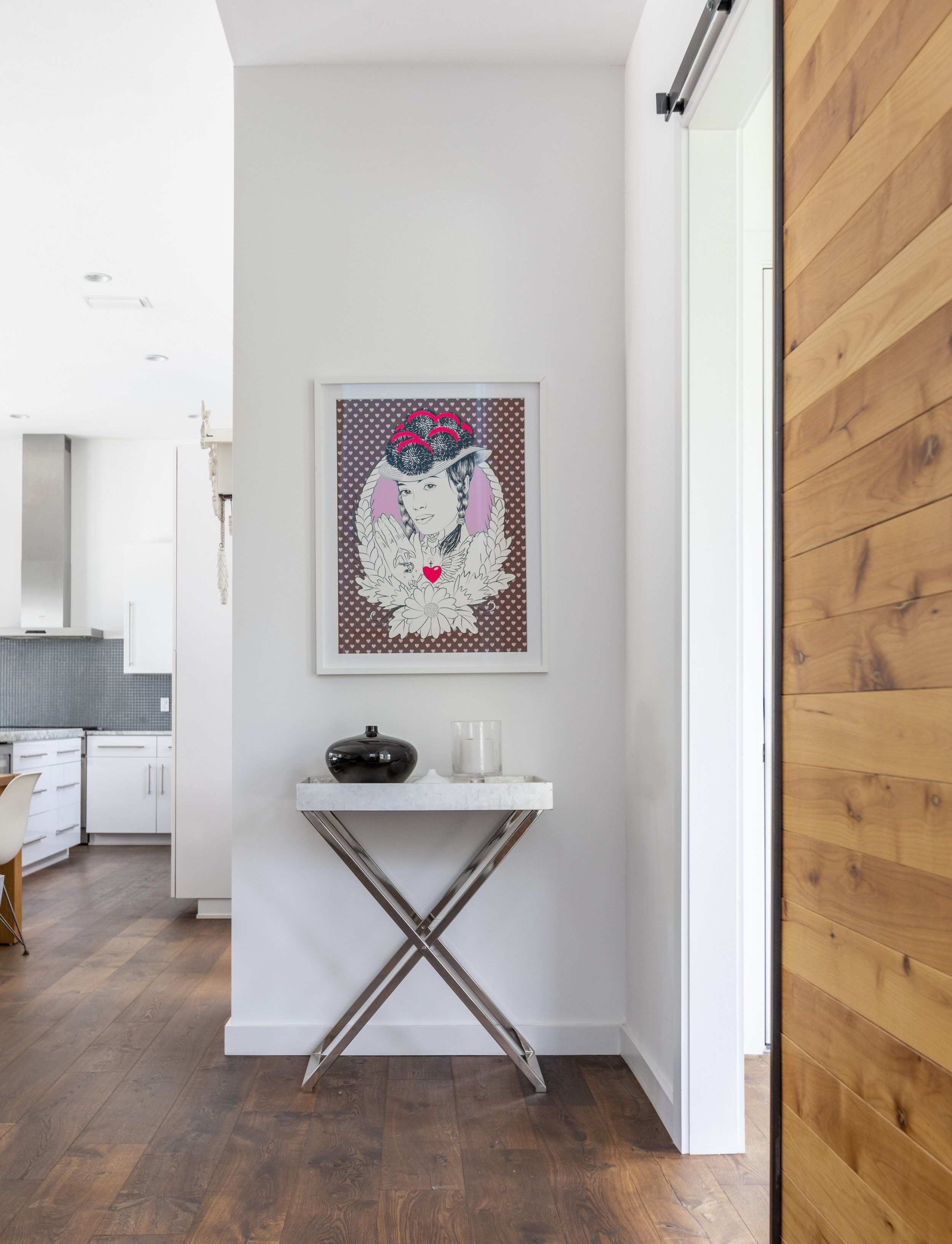 Interior of a modern home with a white wall and wooden flooring. A framed artwork of a woman with braided hair, a hat, and a heart-shaped pendant is hanging on the wall. A small marble table beneath the artwork holds a black vase and a glass. To the 