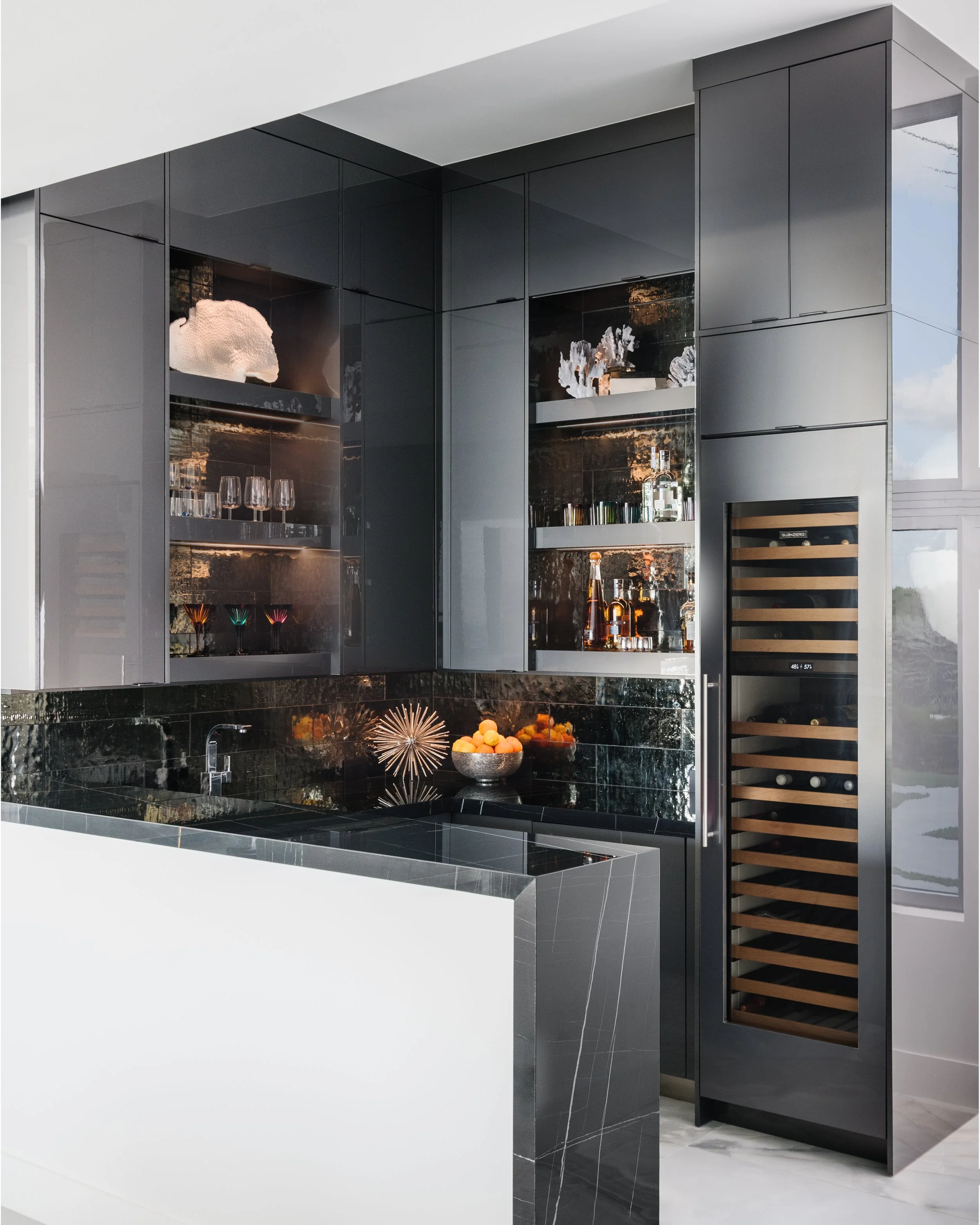 Modern kitchen with black cabinetry, a wine fridge, and black marble countertops and backsplash, with decorative items and a fruit bowl.