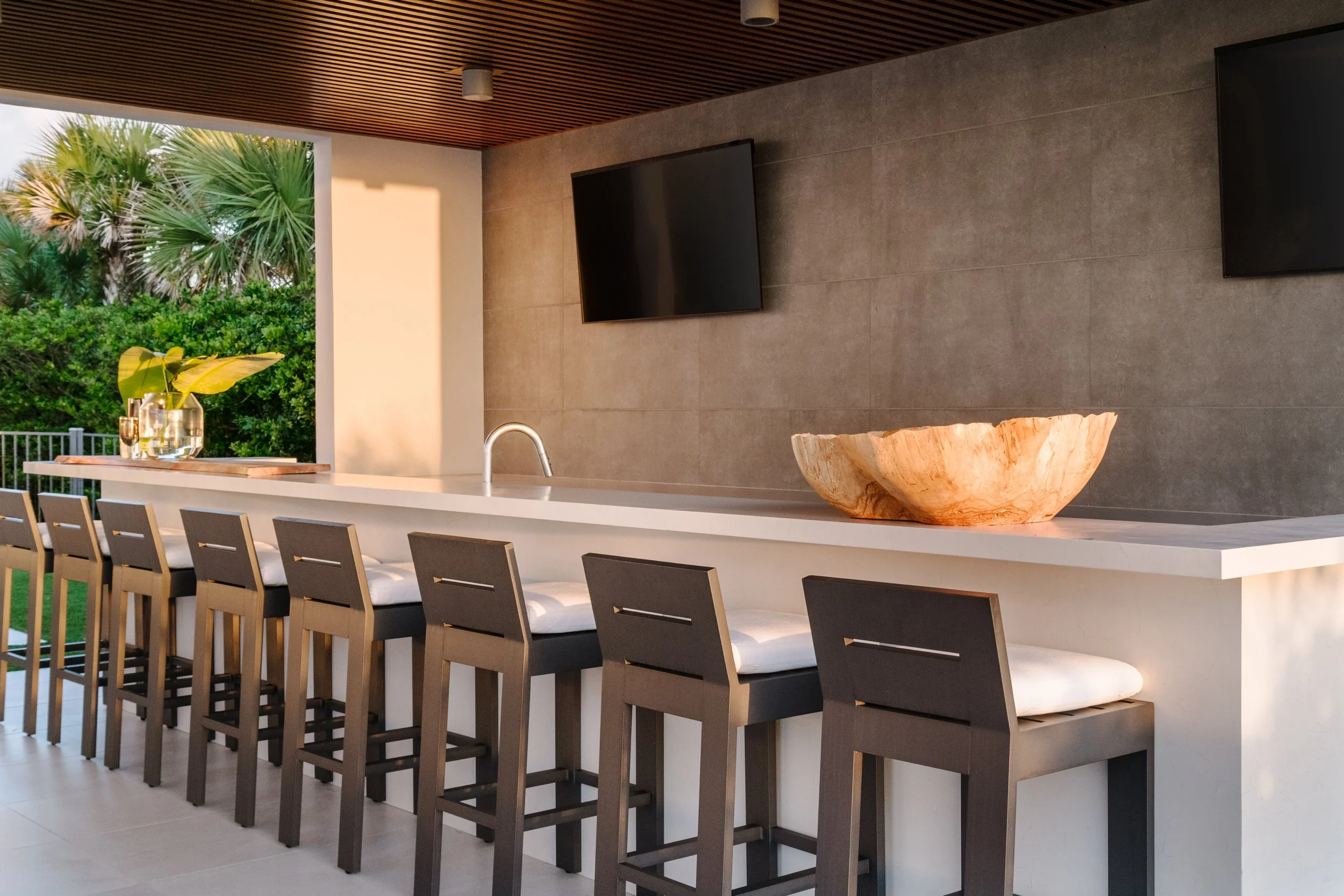 Modern indoor dining area with a long white table, high wooden chairs, and decorative elements including a large wooden bowl and glass vases, overlooking lush green trees outside through an open window.