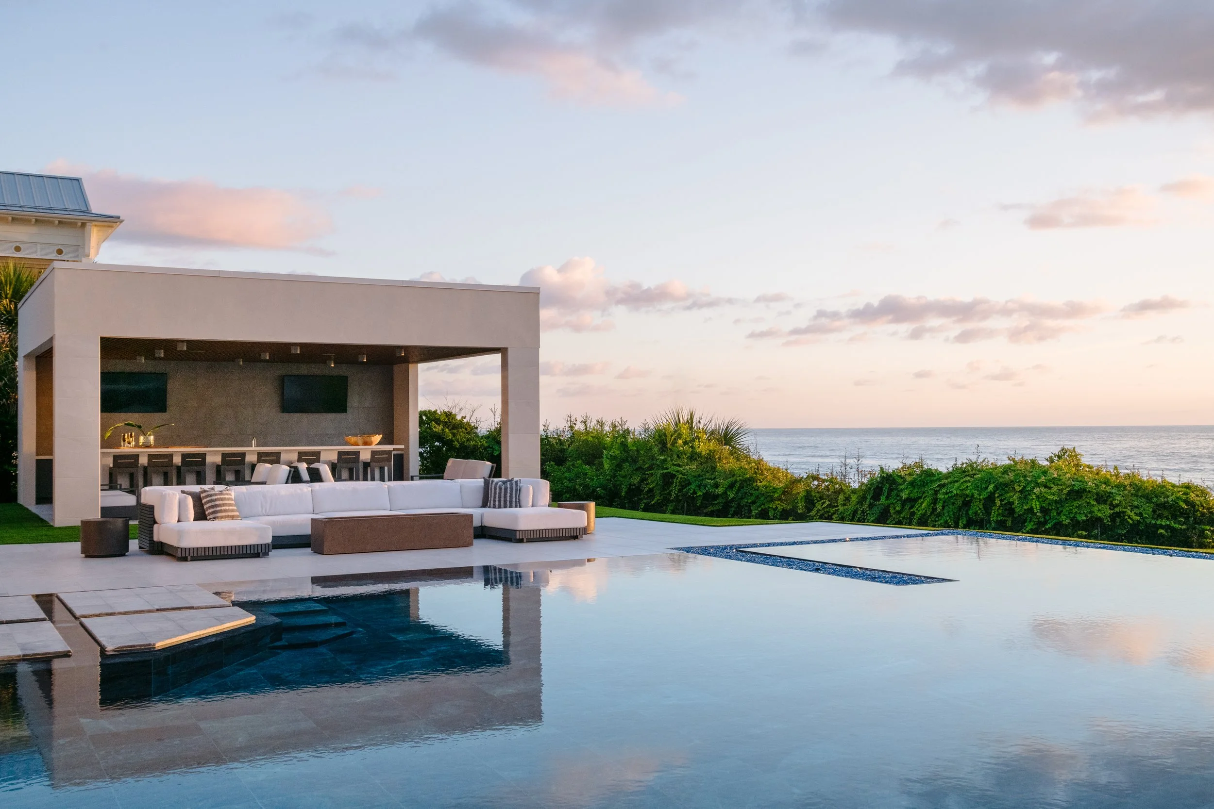 Modern outdoor pool area with white seating, a covered bar or lounge, and ocean view at sunset.