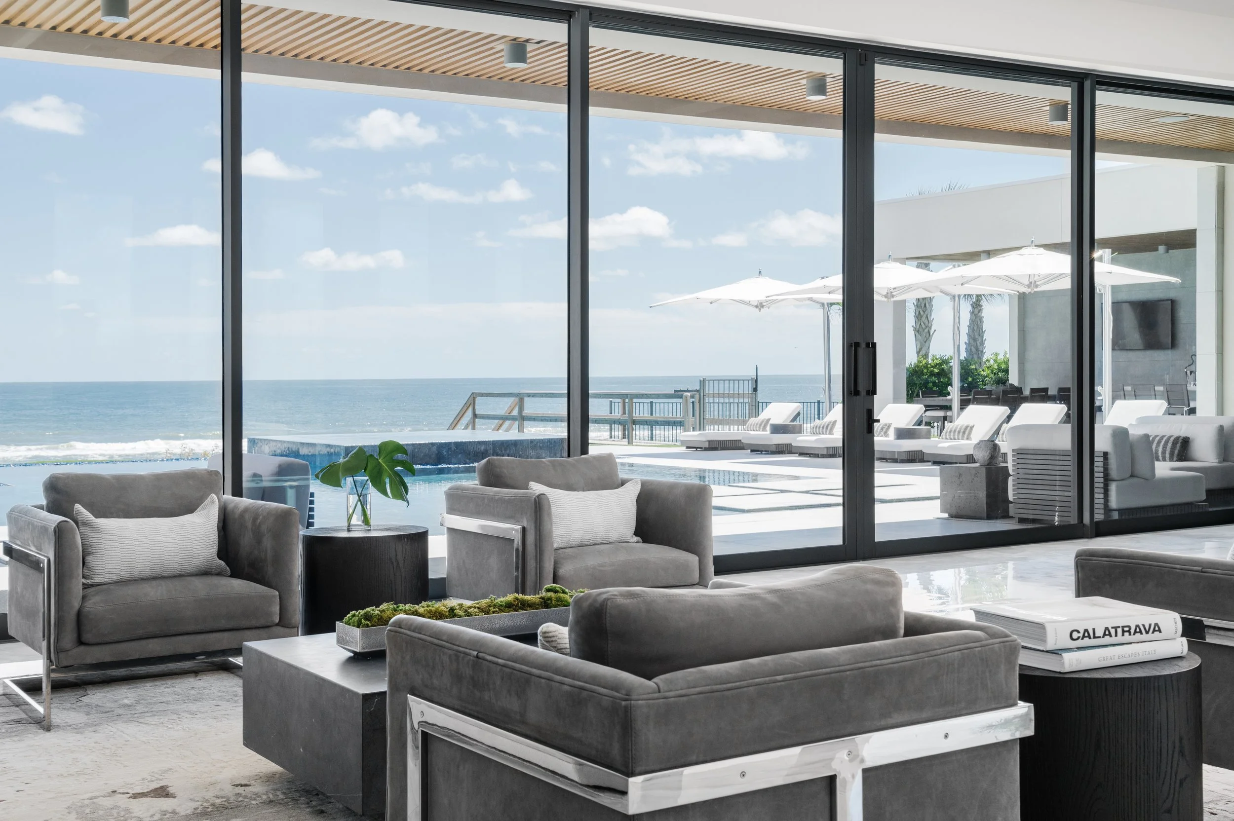 Modern living room with large floor-to-ceiling glass doors showing an ocean view, outdoor lounge chairs, white umbrellas, and a pool area.
