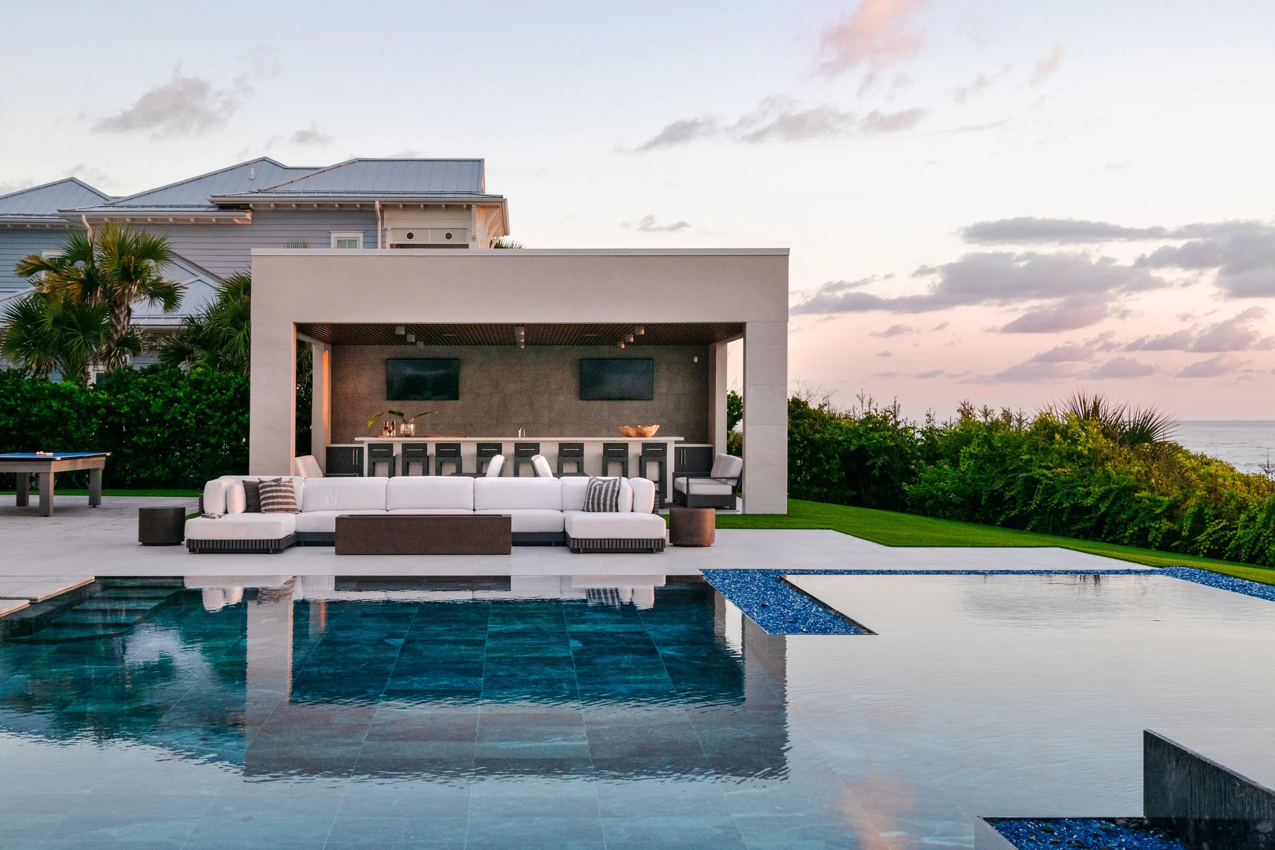Modern outdoor lounge area with a swimming pool, white sectional sofa, bar with stools, greenery, and ocean view at sunset.