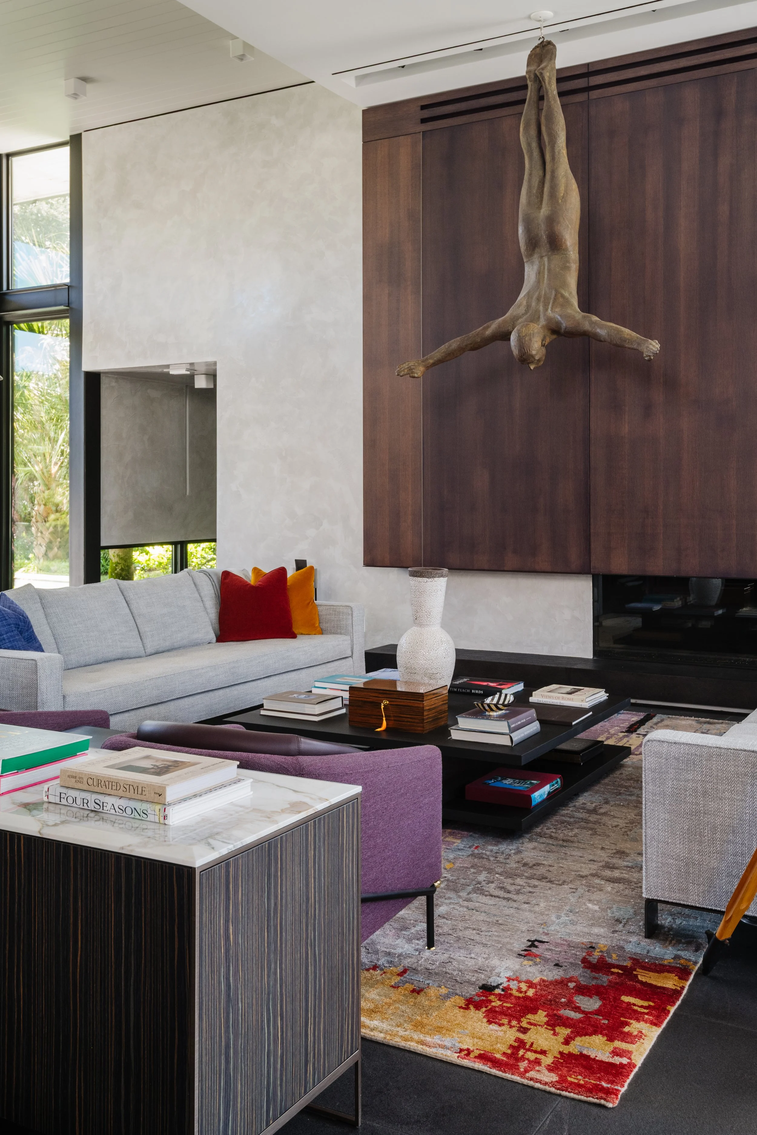 Interior view of a modern living room with a gray sofa, colorful pillows, a black coffee table with books and decorative items, a patterned area rug, a large window with lush greenery outside, and a wooden wall with a suspended sculpture of a human f