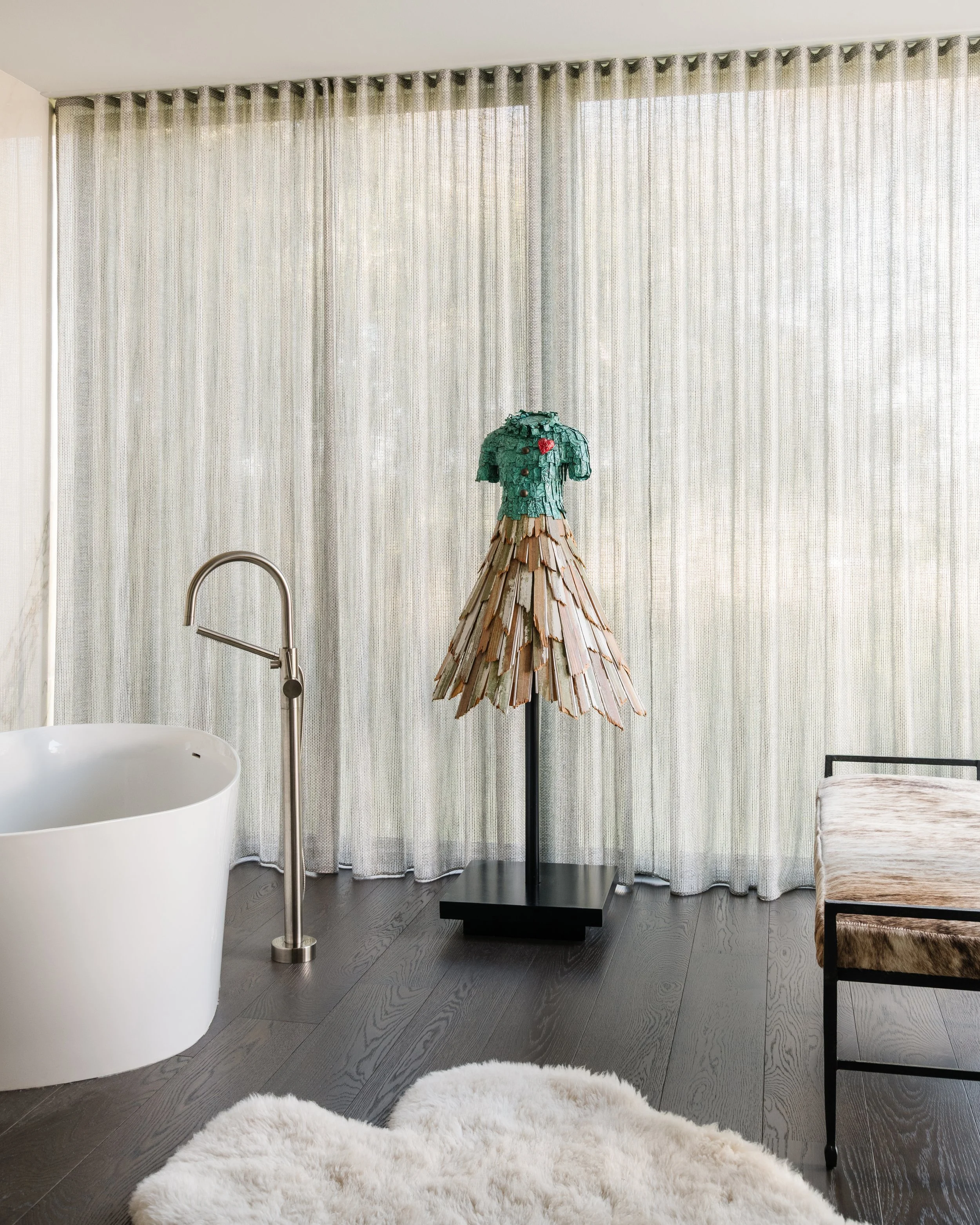 Modern bathroom with a round bathtub, a tall faucet, a decorative dress form with a layered skirt and green top, a light-colored curtain, a black stand, a beige chair, and a white fluffy rug on dark hardwood flooring.