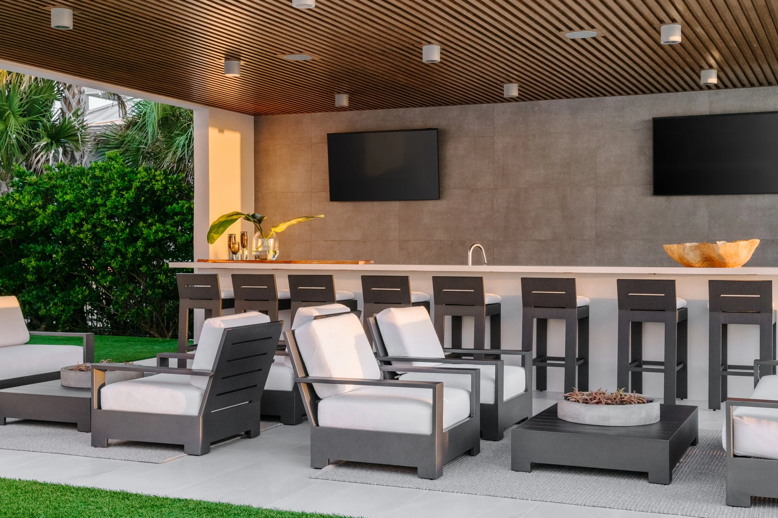 Modern outdoor patio with a bar counter, black chairs with white cushions, sunlit greenery, and two flat-screen TVs mounted on a beige stone wall inside a space with a wooden slat ceiling and recessed lighting.