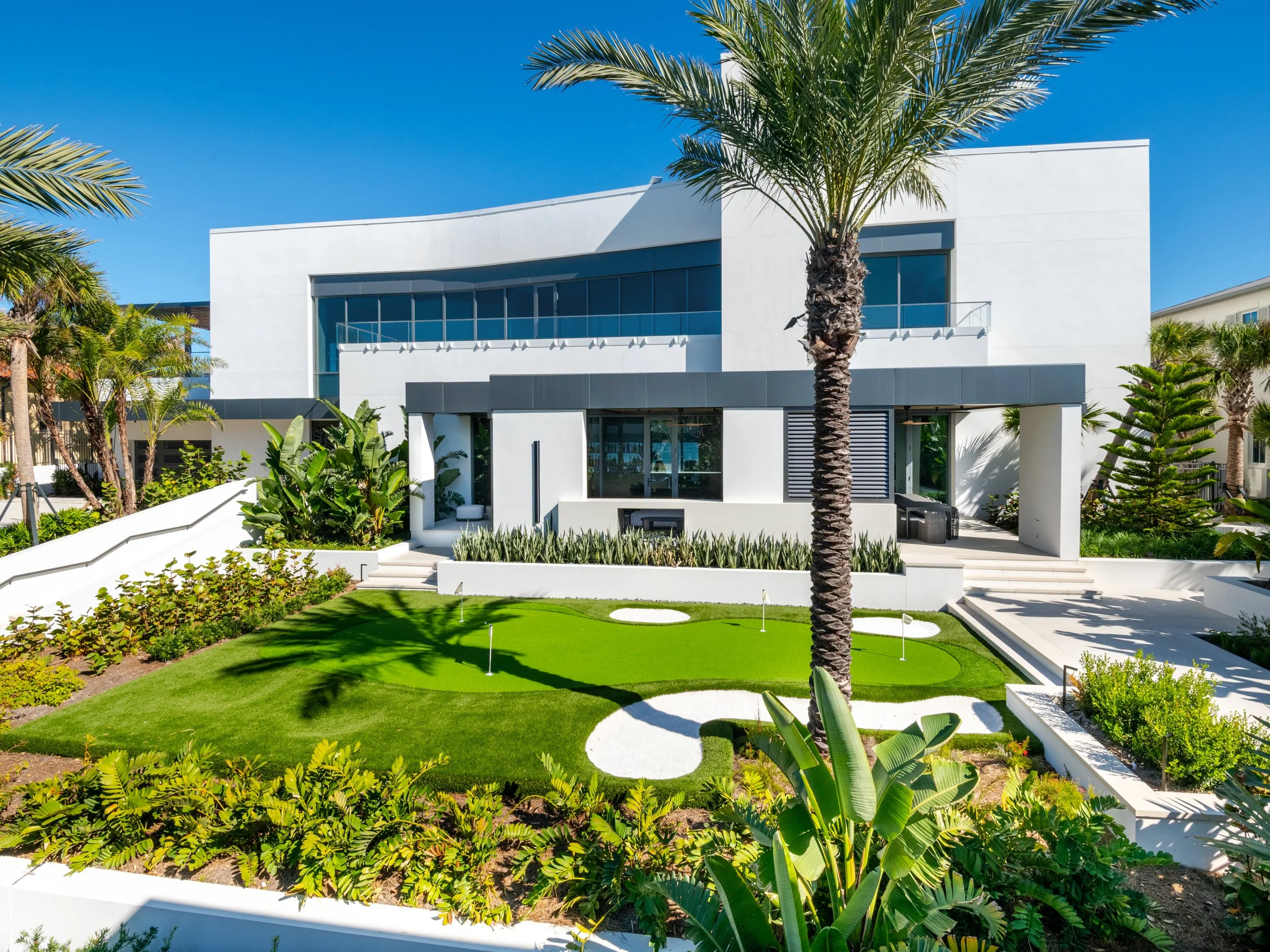 Modern white house with large glass windows, surrounded by lush green landscaping including palm trees and tropical plants, under a clear blue sky.