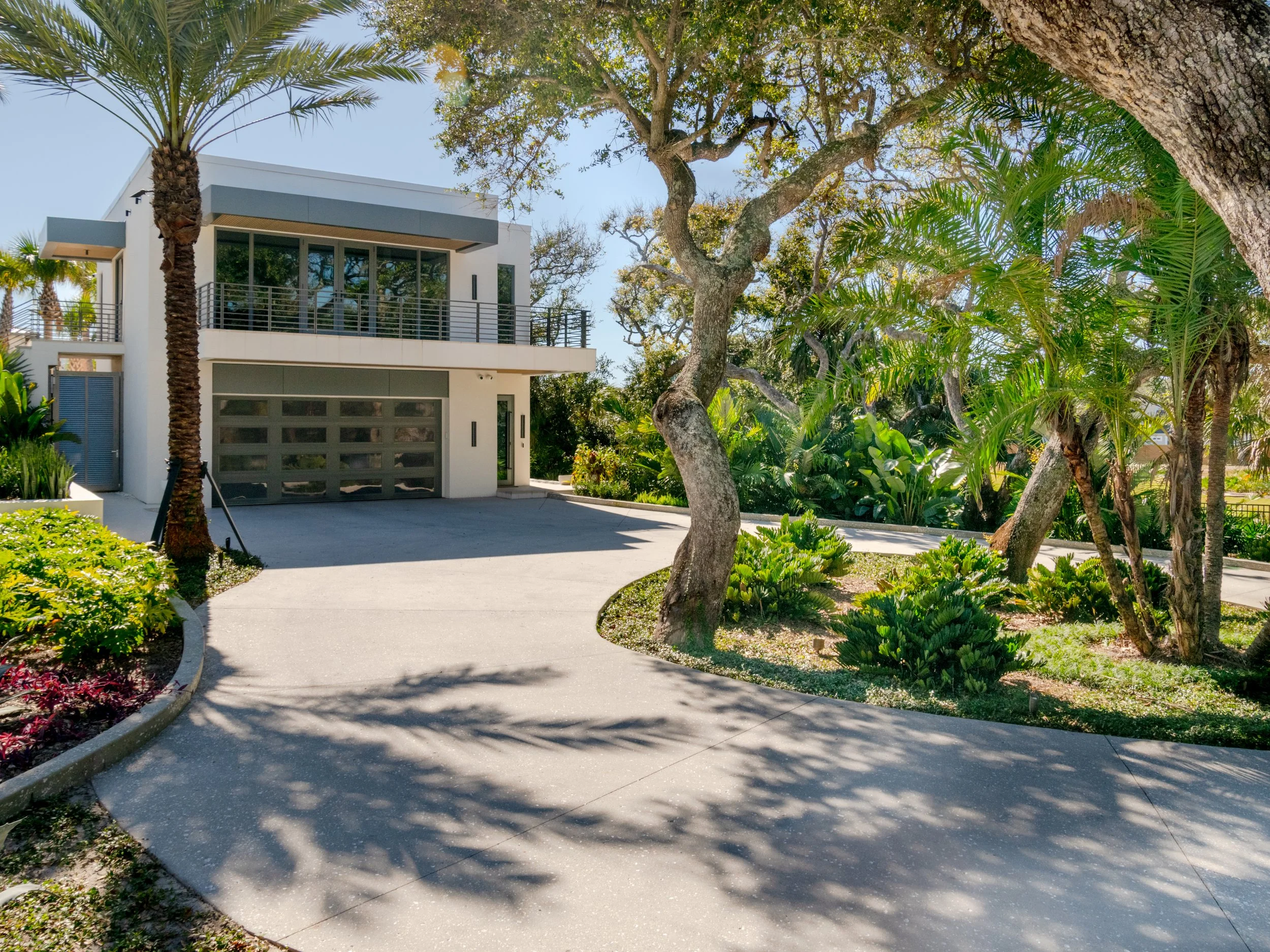 Modern white house with large glass windows and a garage, surrounded by lush green trees and plants on a sunny day.
