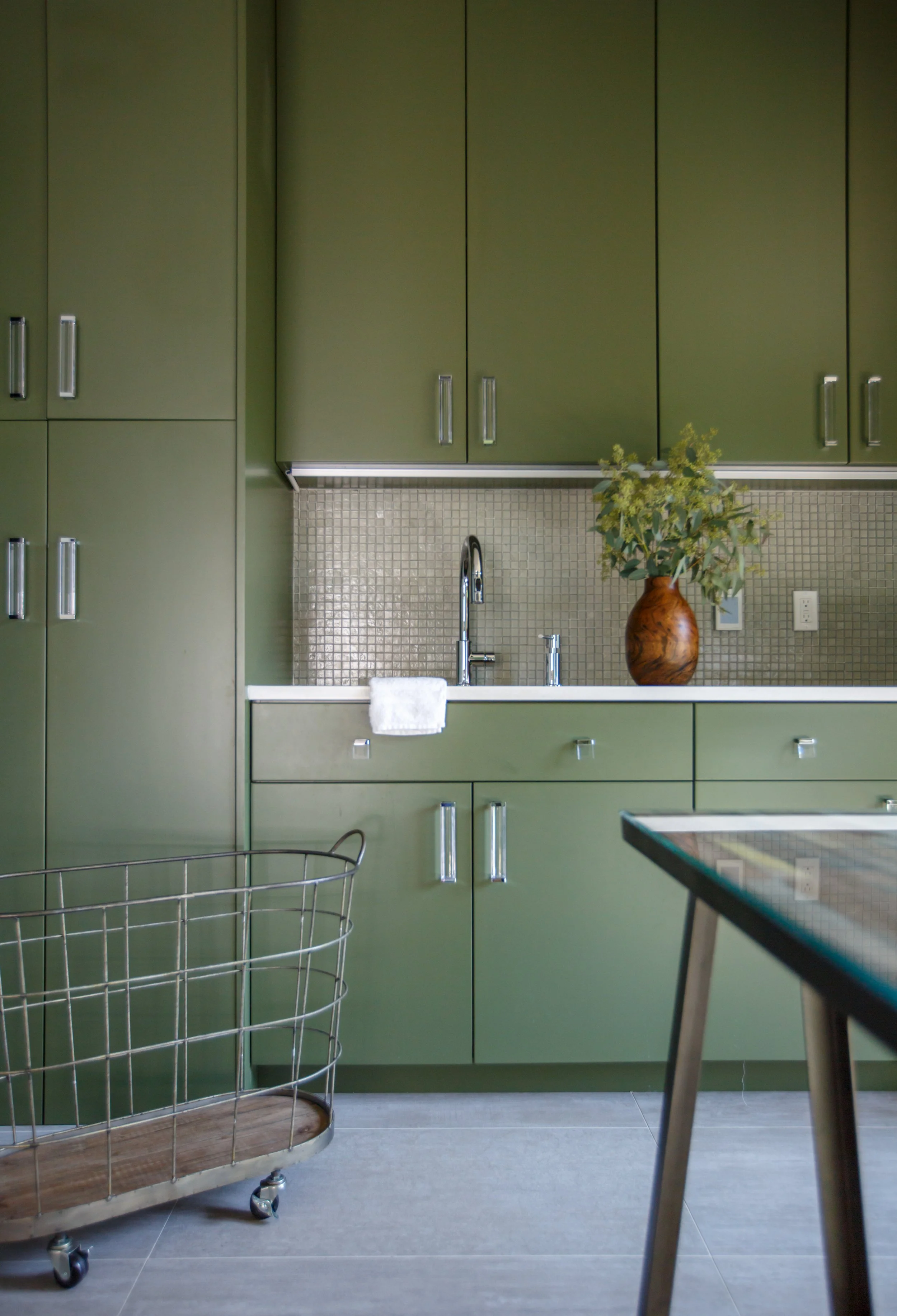 Green kitchen cabinets with silver handles, a beige tiled backsplash, a white countertop with a vase of green foliage, a kitchen sink with a chrome faucet, a white towel hanging, a metal wire chair with wooden armrest, and a glass table in the foregr