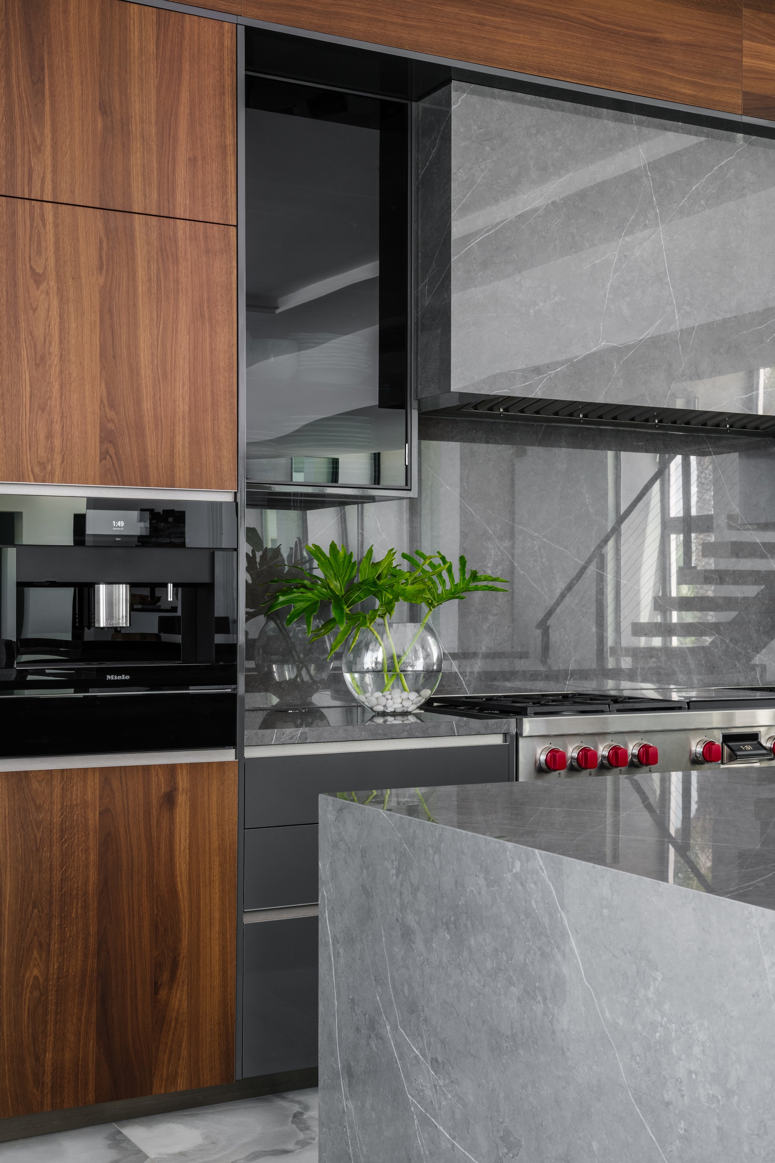 Modern kitchen with wood cabinetry, gray marble countertops and backsplash, a built-in oven, a potted plant in a clear glass bowl, and a stove with red knobs.