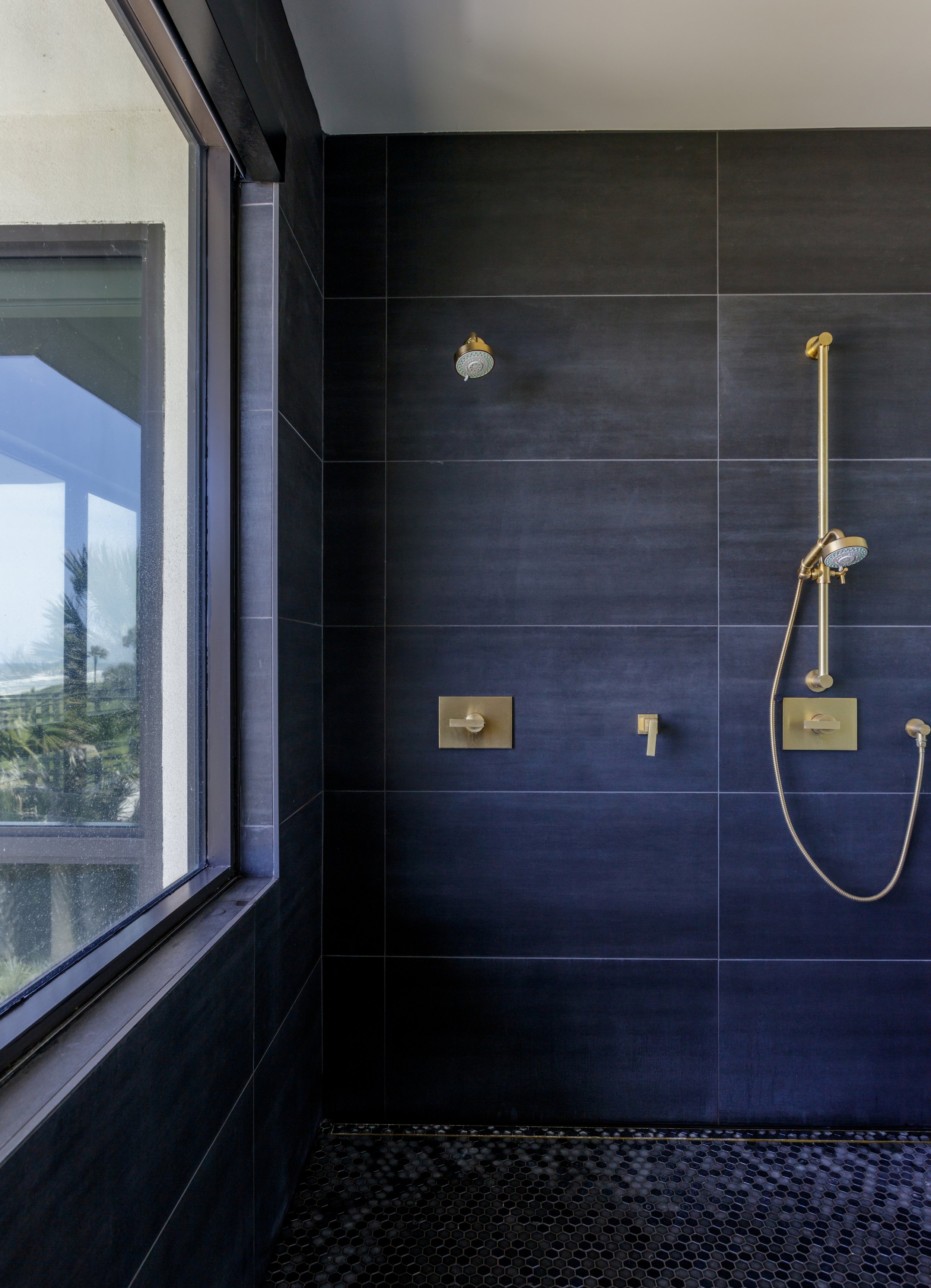 Inside a modern shower area with black tiled walls, a brass showerhead on a horizontal bar, and a window letting in natural light.