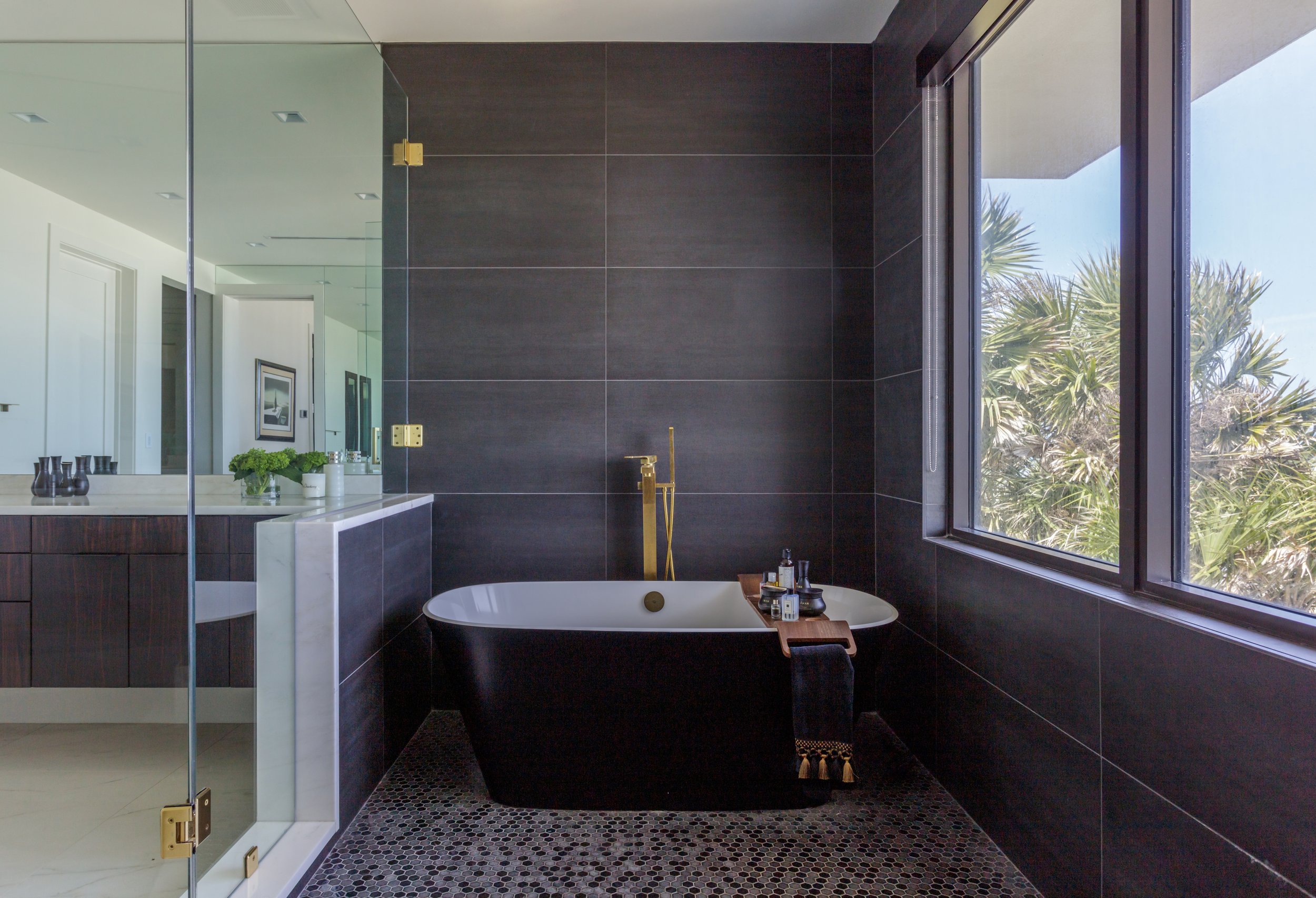 Modern bathroom with black tiled walls, a freestanding bathtub, and a large window showing trees outside.