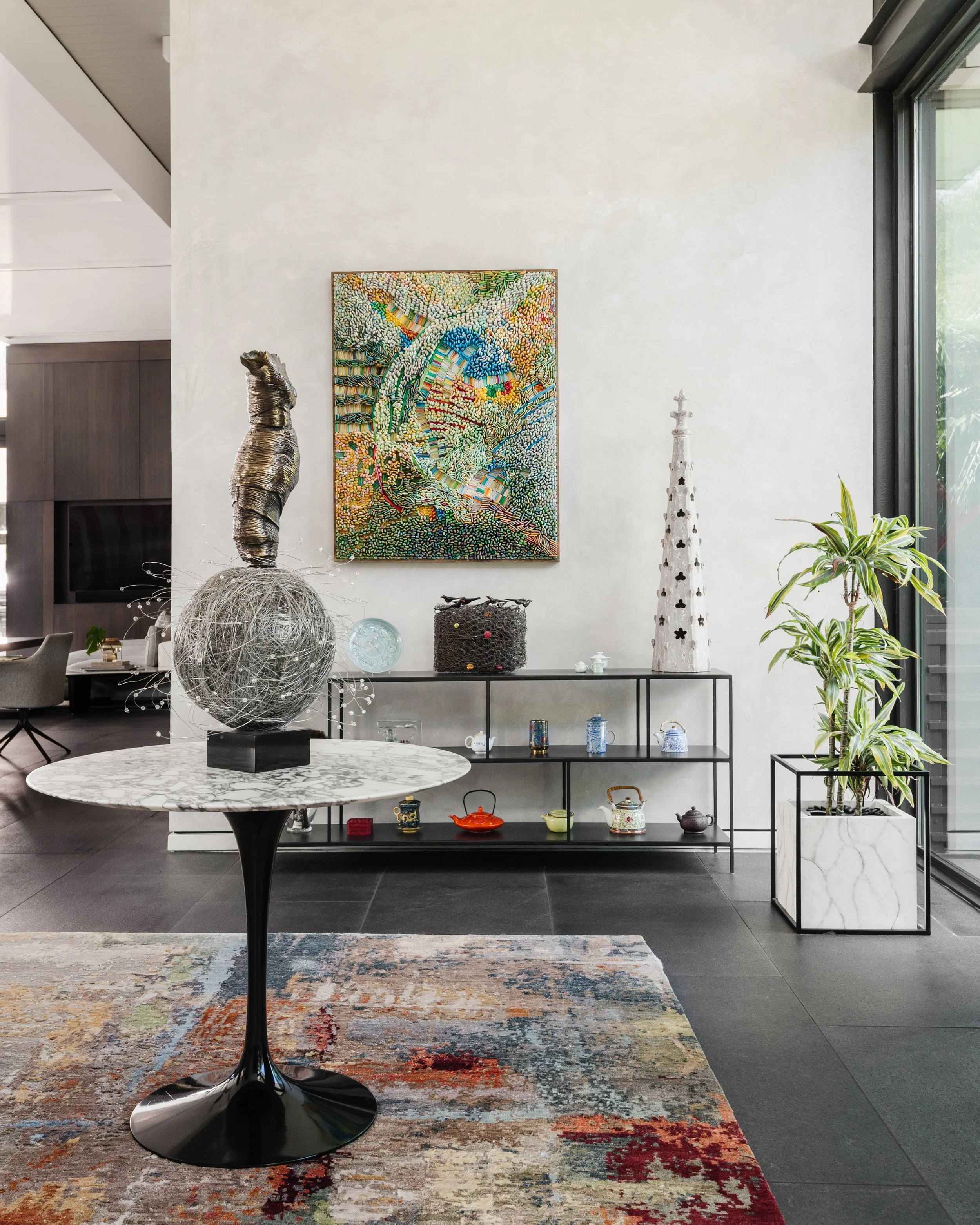 Modern interior with abstract painting on white wall, decorative items including a metal sculpture, a white marble-topped table with a black pedestal base, and various vases and plants, all on a colorful distressed rug.