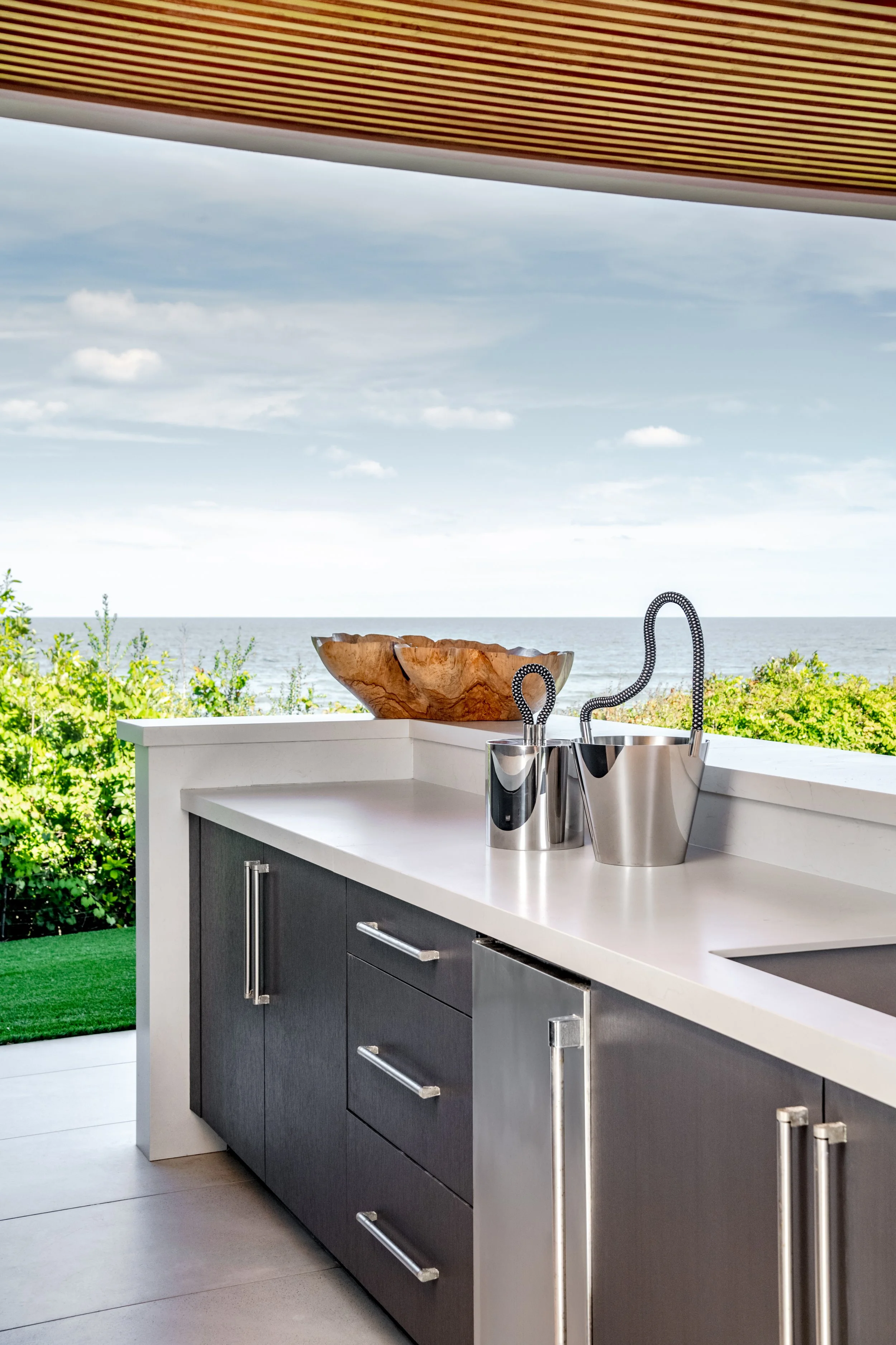 Outdoor kitchen counter with a wooden bowl and a modern stainless steel sink with flexible hose, overlooking the ocean and greenery under a wooden pergola.