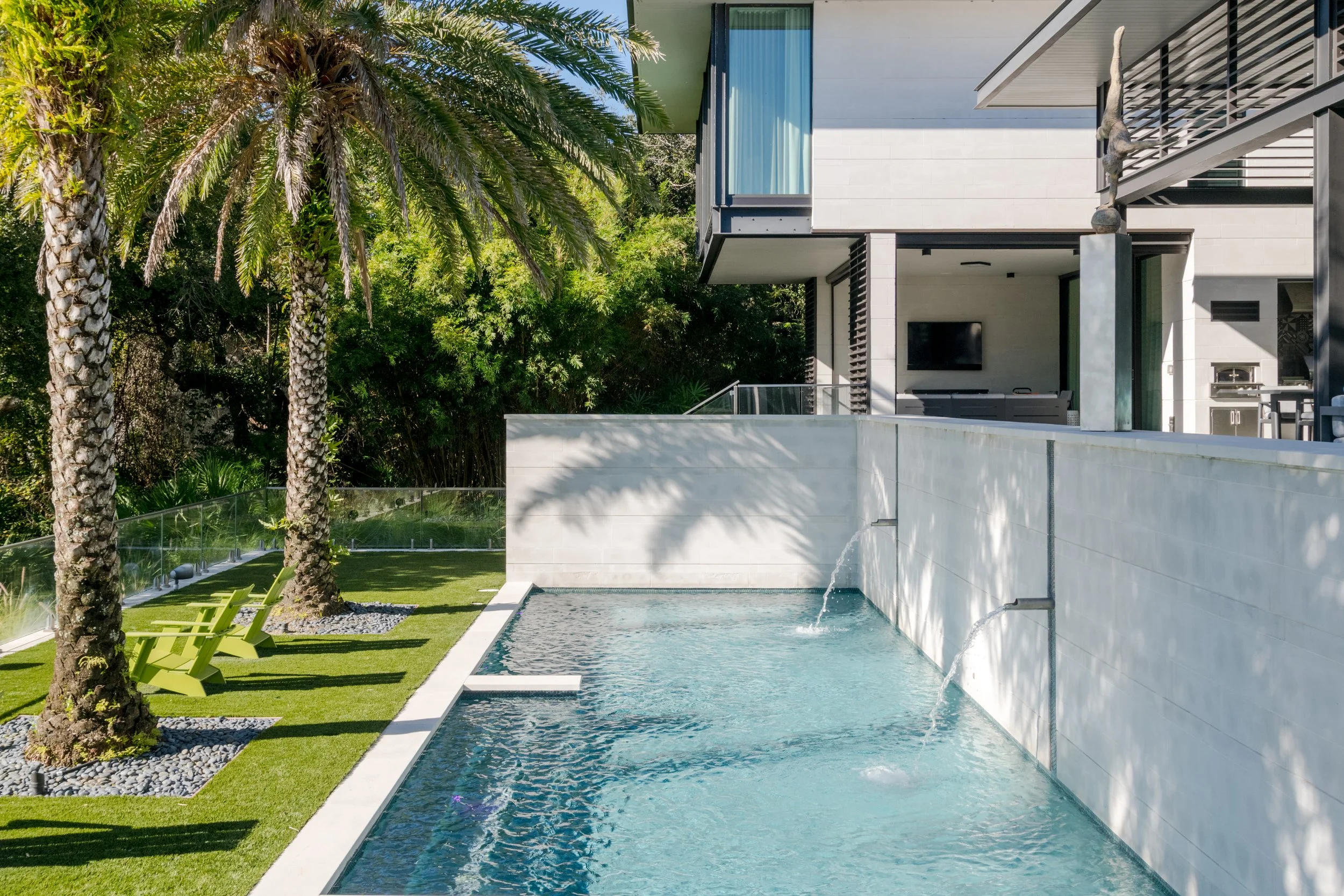 Modern house with a backyard pool, tall palm trees, outdoor green chairs, and a lush green garden area.