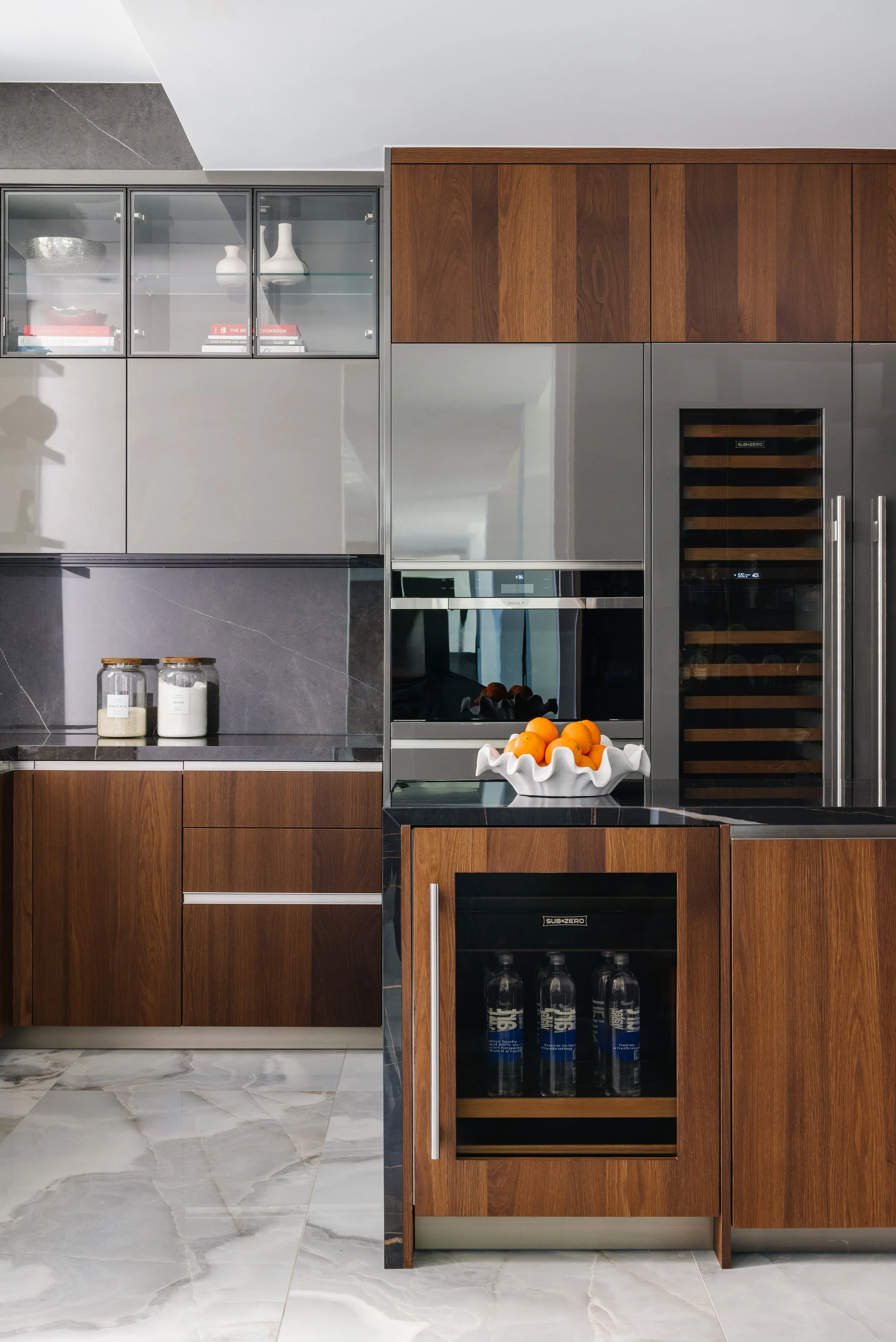 Modern kitchen with wooden cabinets, gray metallic appliances, marble flooring, and a bowl of oranges on the countertop.