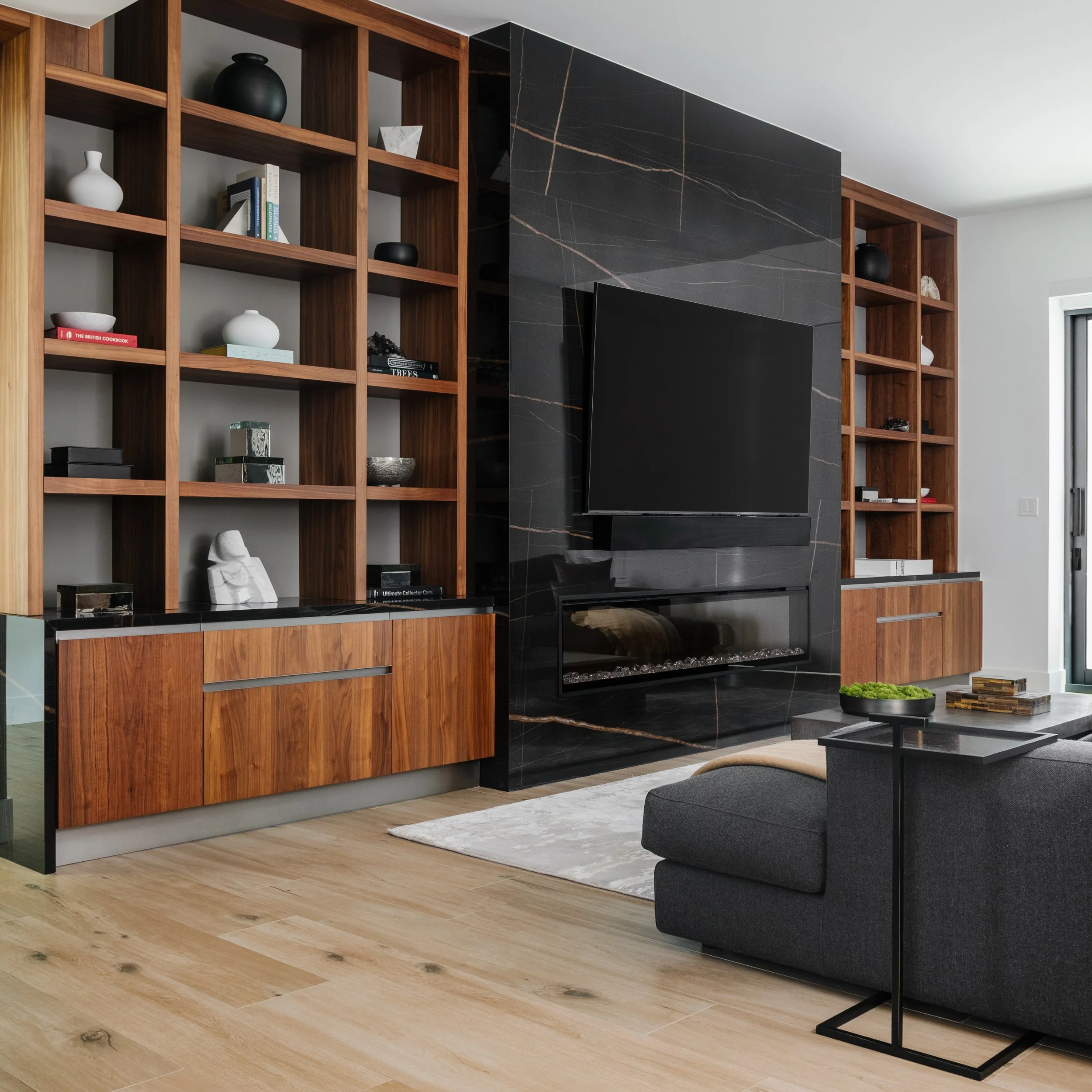 A modern living room with wooden bookshelves, a black marble fireplace, a gray sofa, a black side table with a potted plant, and hardwood flooring.