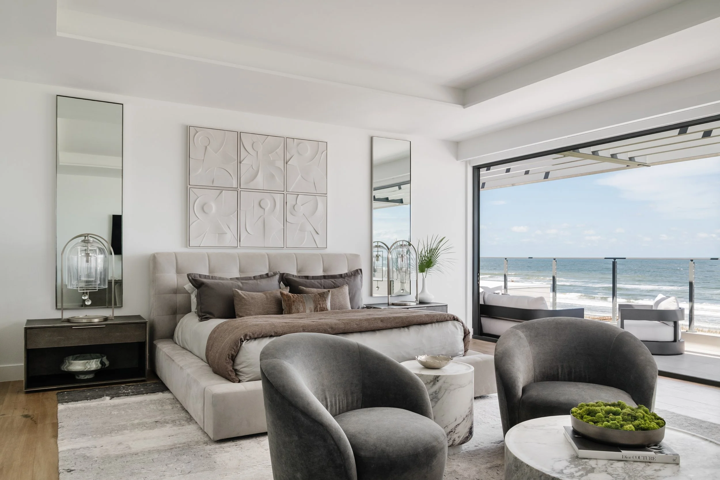 A modern, minimalist bedroom with a large window/door opening to a beach view, white walls, a light-colored upholstered bed with decorative pillows, and nearby mirrors and side tables. There is a marble-topped table with a green moss decoration.