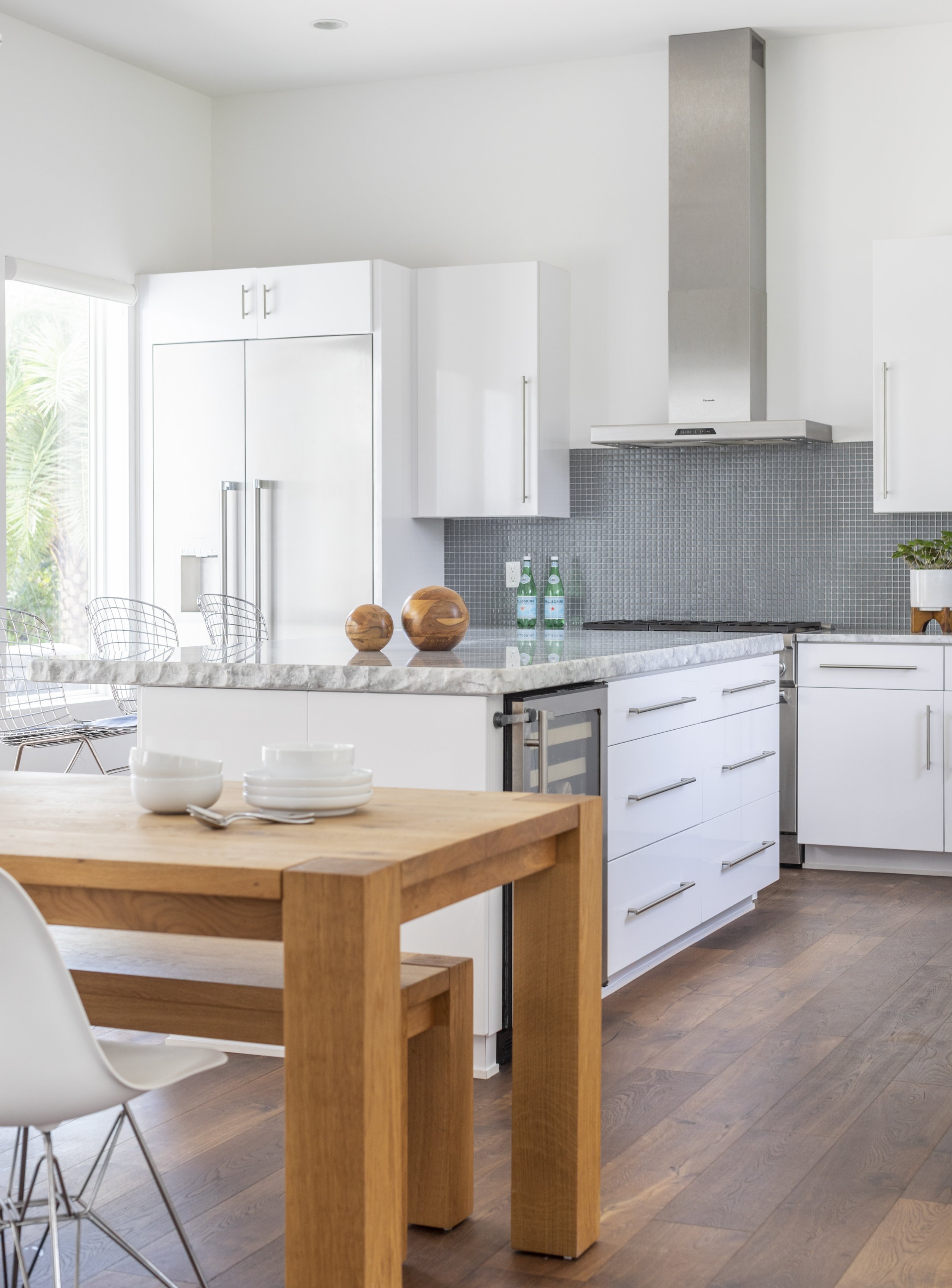 Modern kitchen with white cabinets, gray backsplash, stainless steel range hood, and wooden dining table with white bowls and silverware.