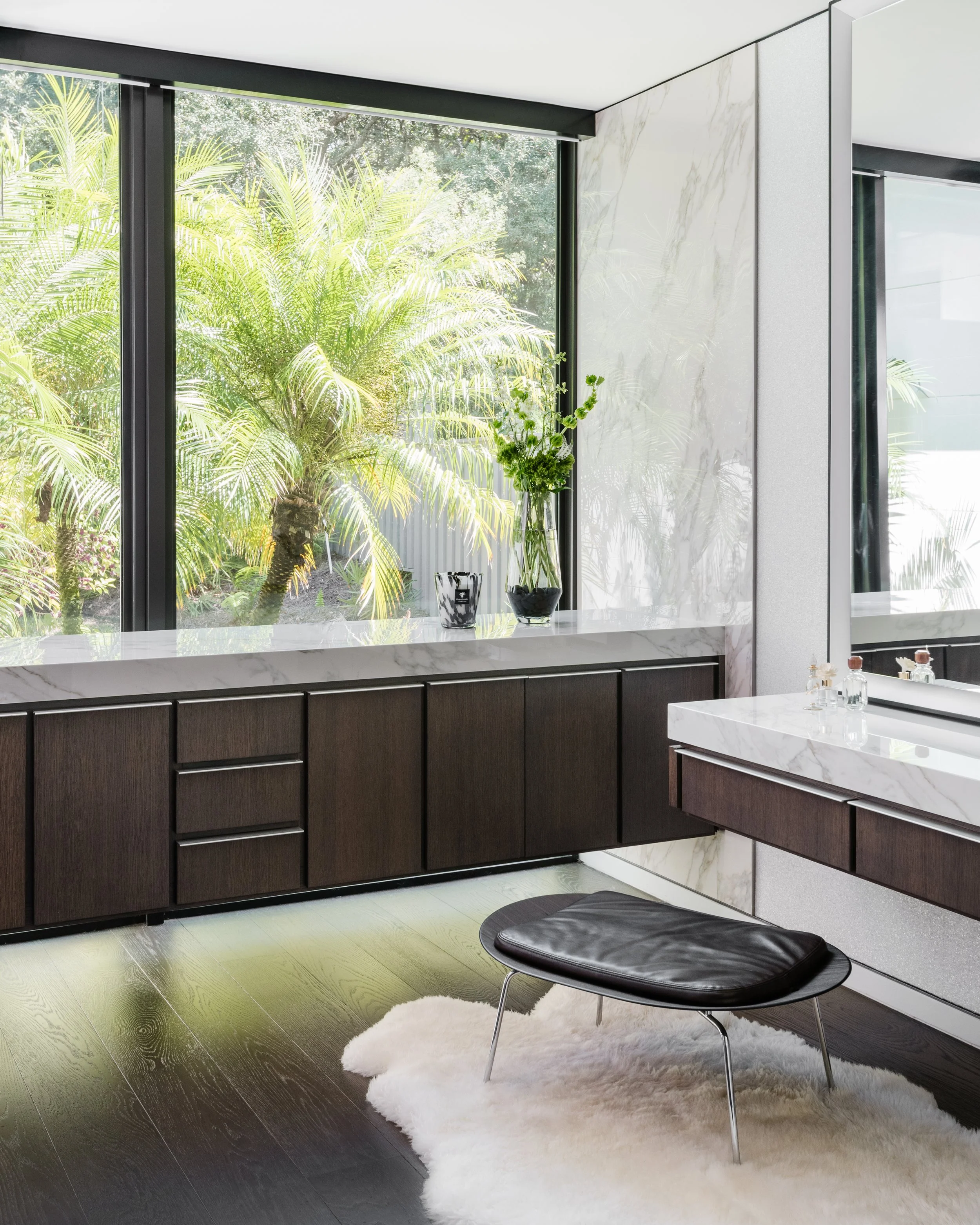 Modern kitchen with black cabinets, marble countertops, large window with greenery outside, small plant on counter, black chair with white rug nearby.
