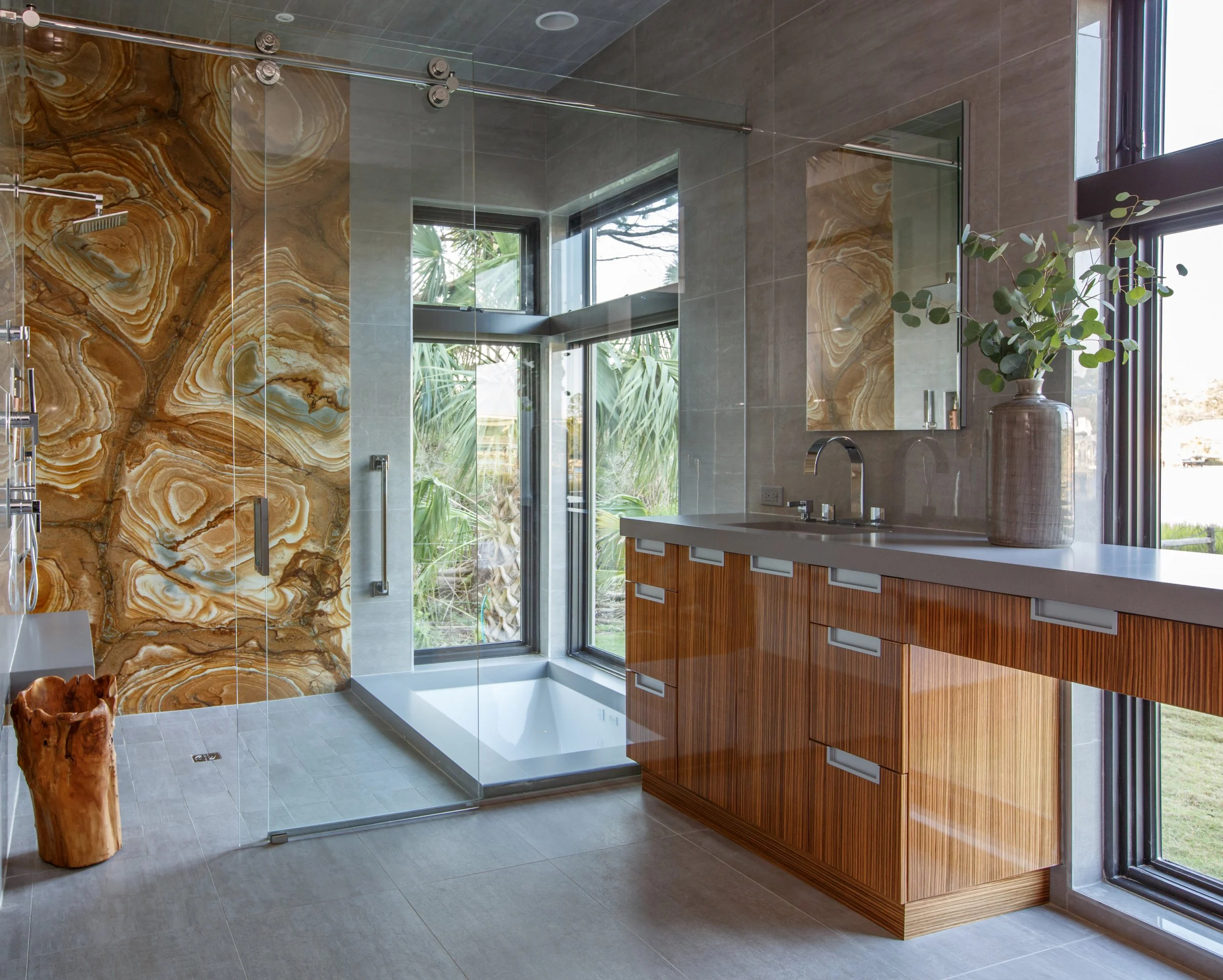 Modern bathroom with a walk-in shower with glass door, brown and beige marble wall, large window showing greenery outside, wooden vanity with quartz countertop, oval mirror, and a potted plant.