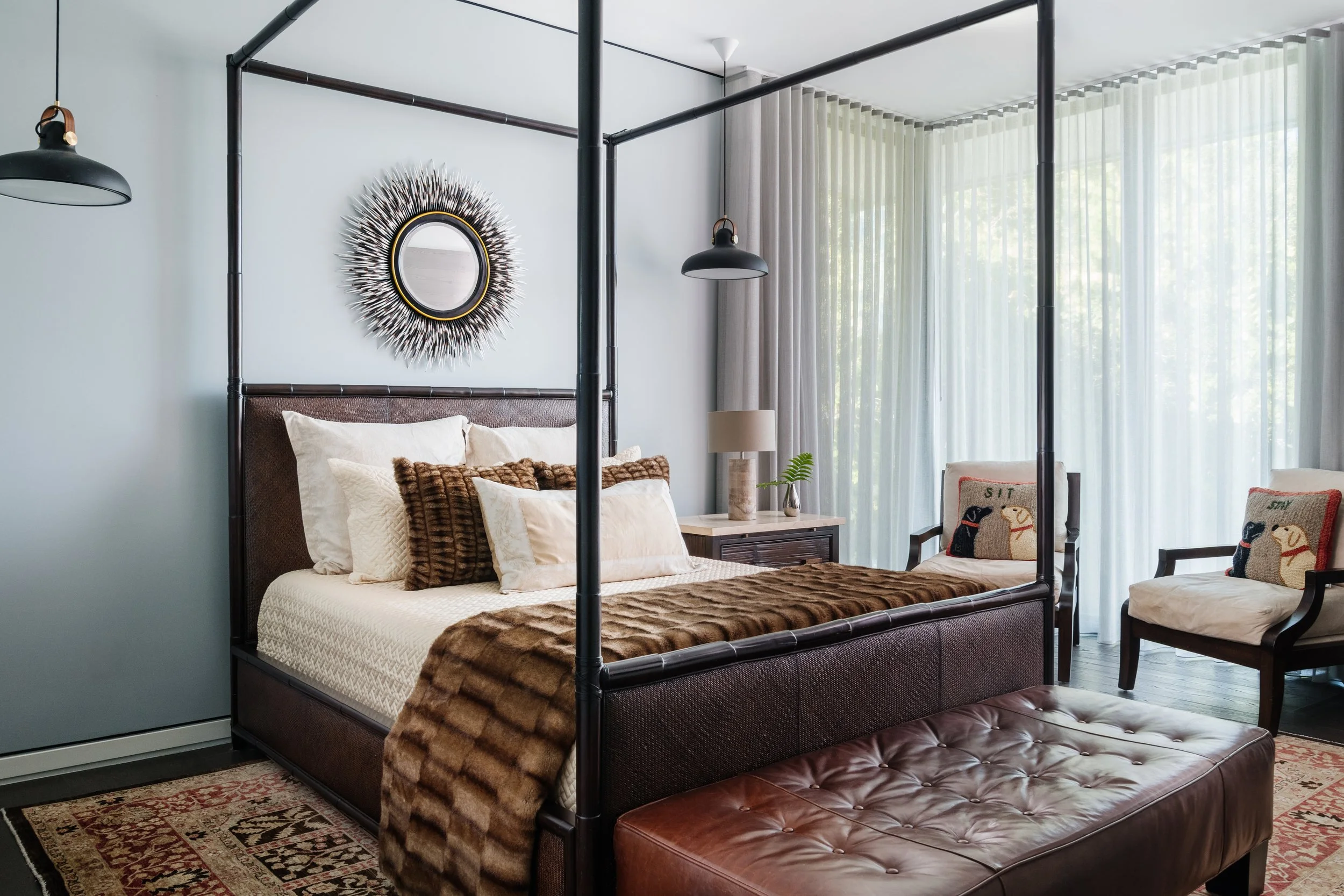 A bedroom with a four-poster bed, pillows, a wall mirror, lamps, and chairs with decorative pillows, near large windows with sheer curtains.