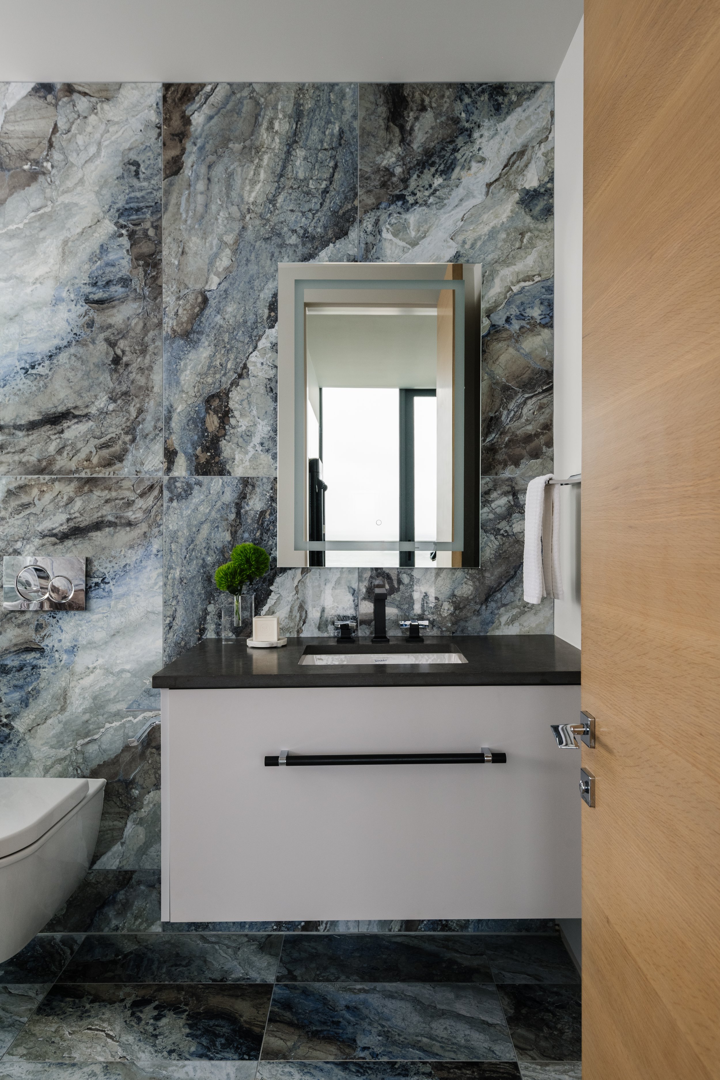 Modern bathroom with marble walls, a mirror above a black countertop with a sink, decorative plant, and towel rack, with wooden door to the right.