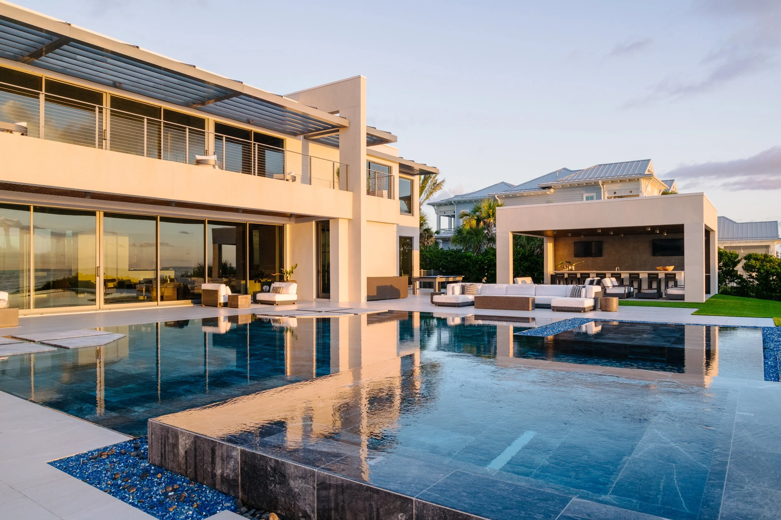 Modern beachfront house with multiple levels, large glass windows, outdoor seating, and a swimming pool reflecting the sky and building, during sunset.