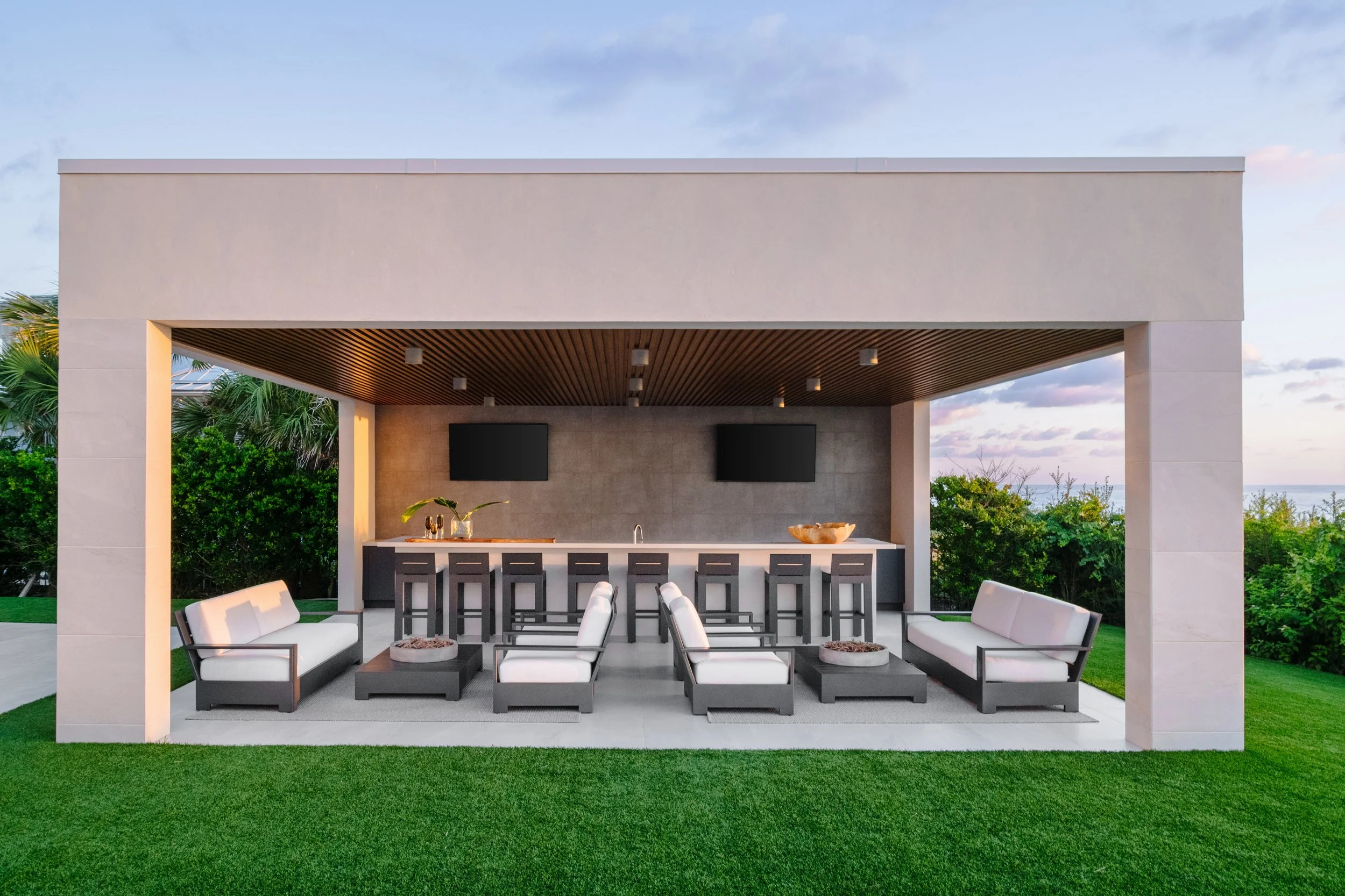Modern outdoor living space with a covered bar area, seating, and lush greenery, overlooking a body of water with a partly cloudy sky.
