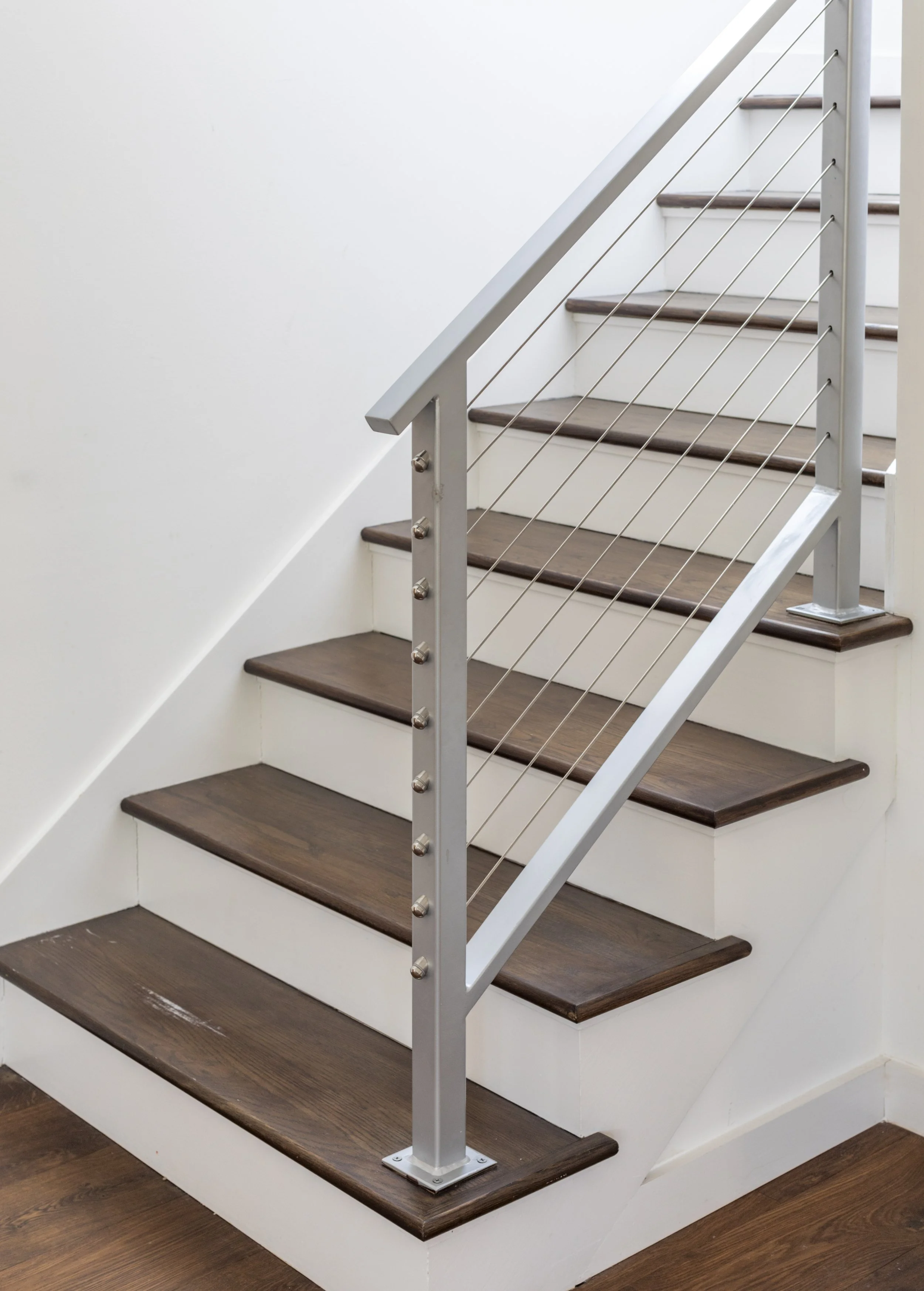 Wooden staircase with white metal handrail and horizontal metal wires, walls painted white and wooden floor.