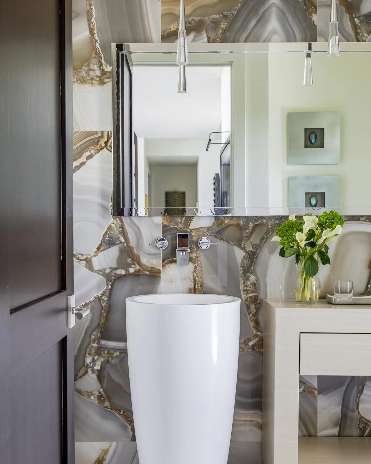 Modern bathroom with marble accent wall, rectangular mirror, white free-standing bathtub, beige vanity with a vase of white calla lilies, wall art, and pendant lighting.