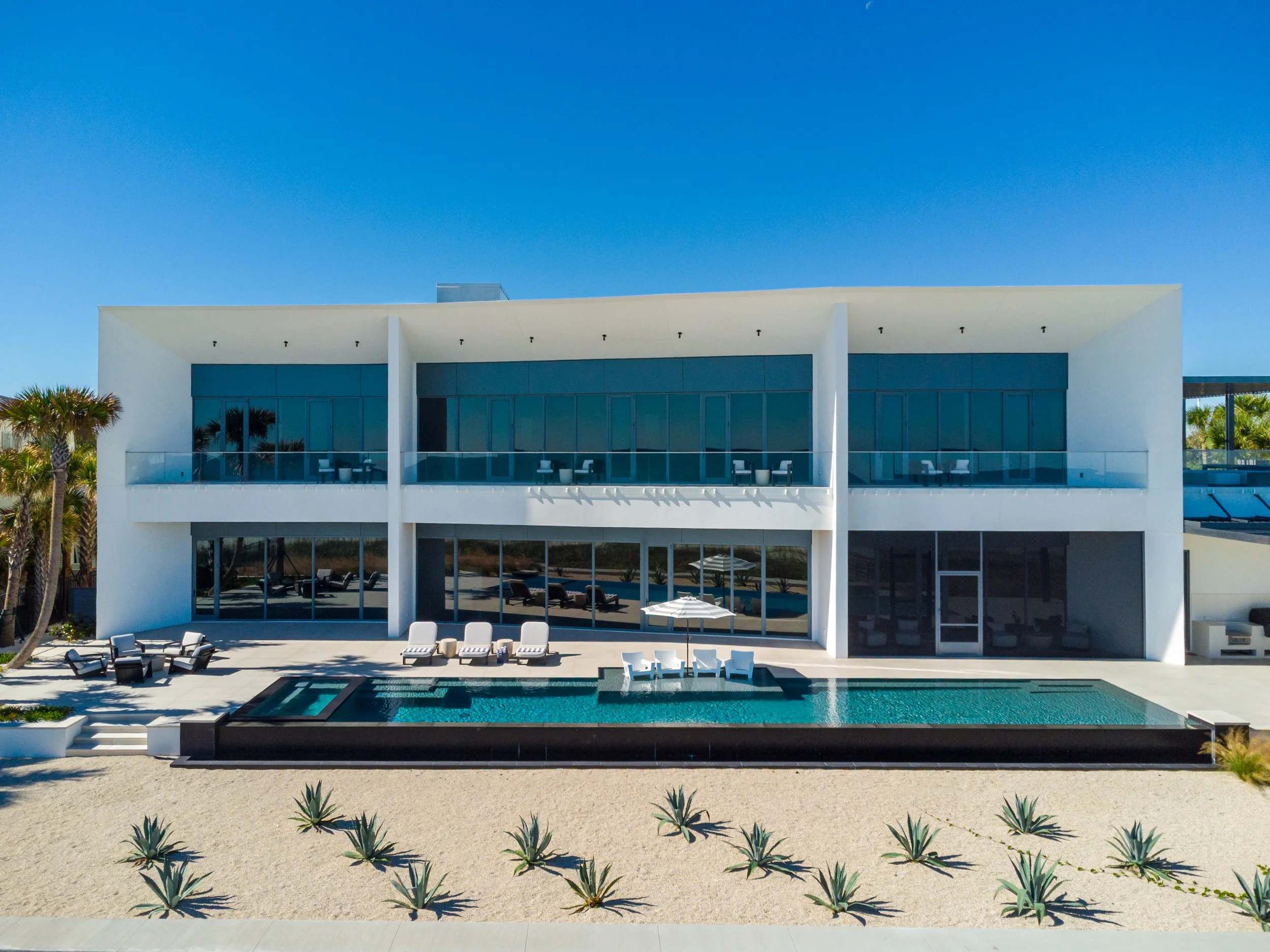 Modern white house with large glass windows, outdoor pool, and lounge chairs, surrounded by desert plants and palm trees, under a clear blue sky.