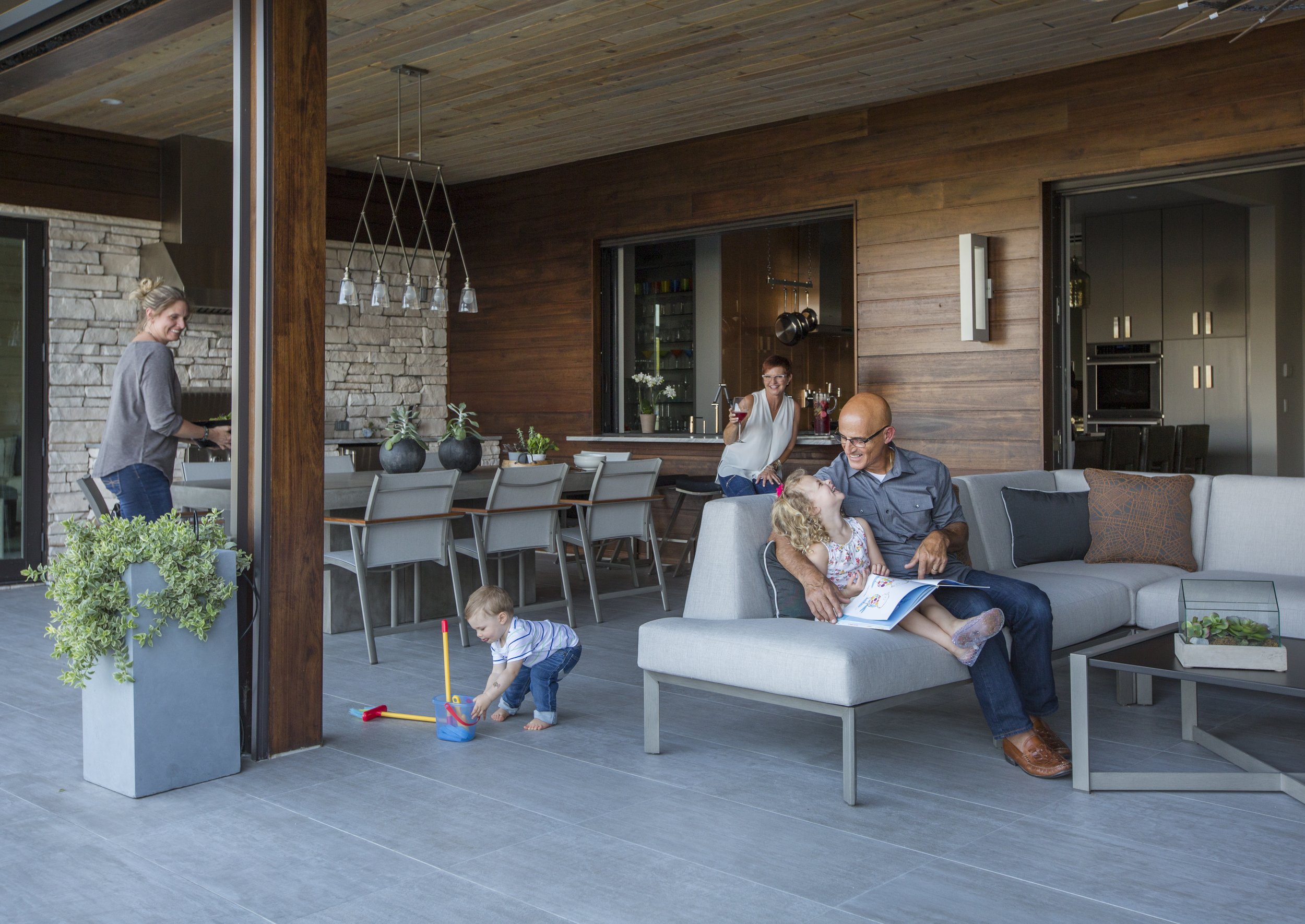 A family enjoying time on their outdoor patio with a modern wood and stone design, children playing on the ground, and adults relaxing and chatting.