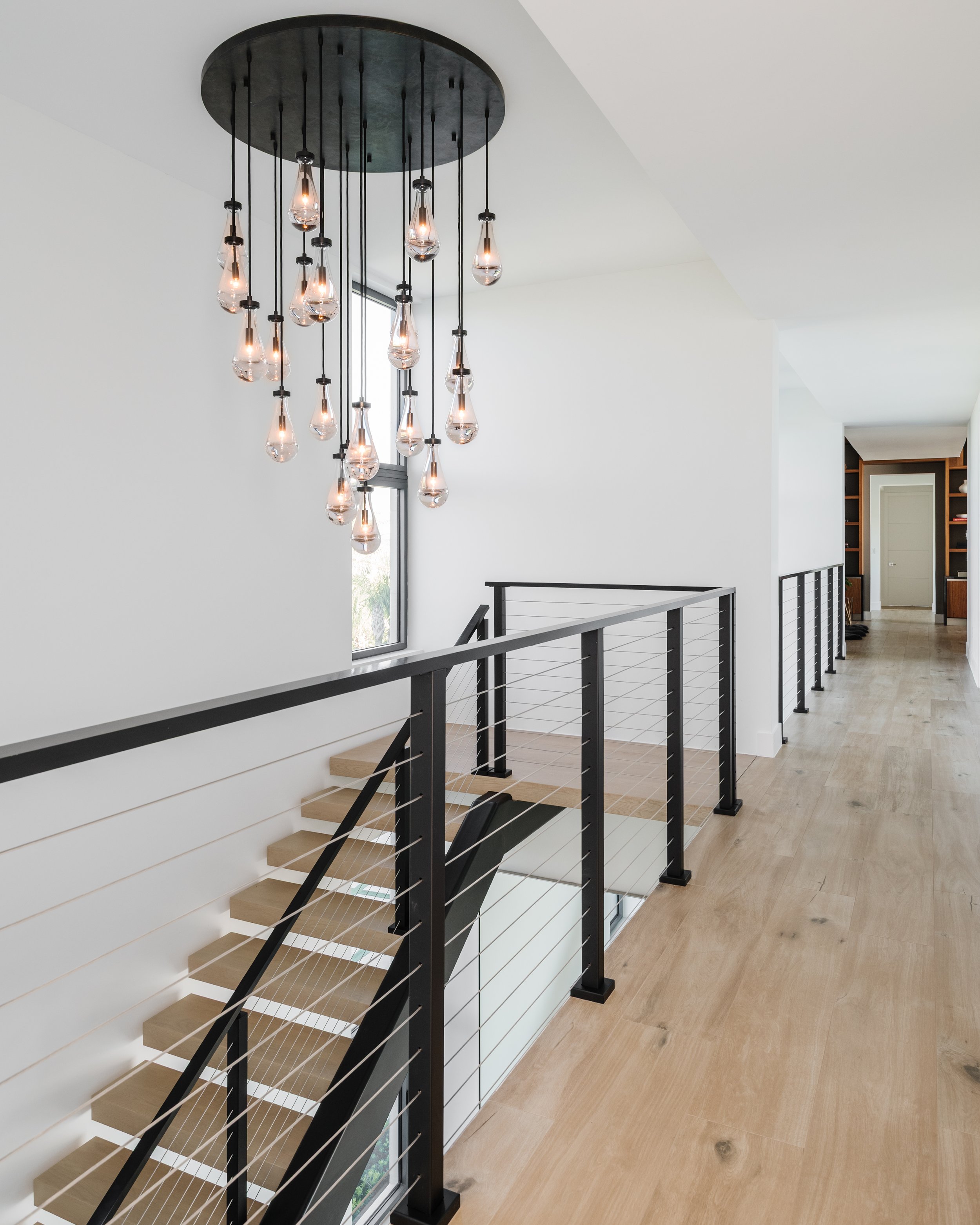 Modern staircase with black metal railing, wooden steps, and a hanging chandelier with multiple exposed bulbs inside a bright, white wall interior.