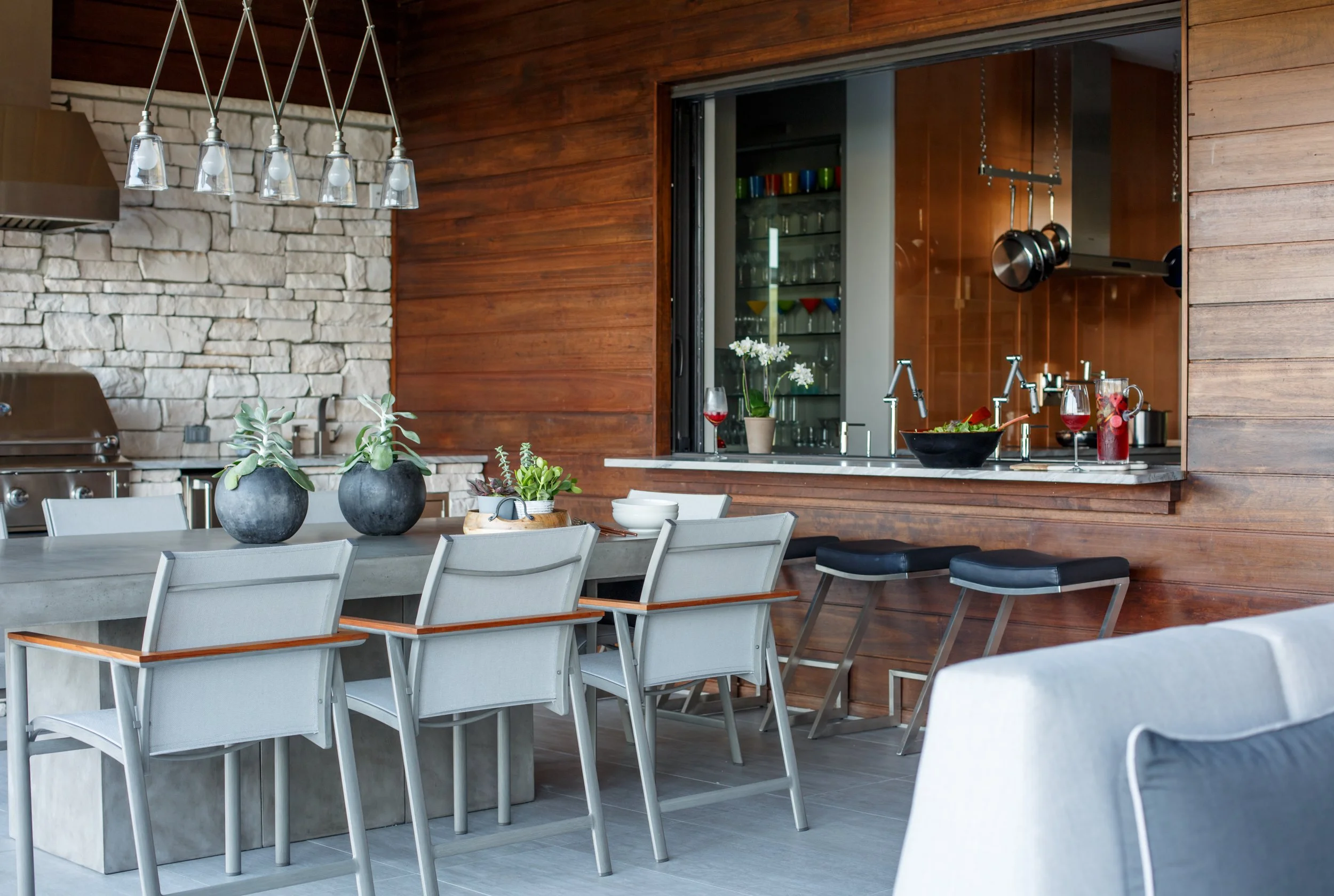 Modern kitchen with wooden and stone walls, white dining table with beige chairs, decorative plants, and open view to a kitchen area with glassware and hanging pots.