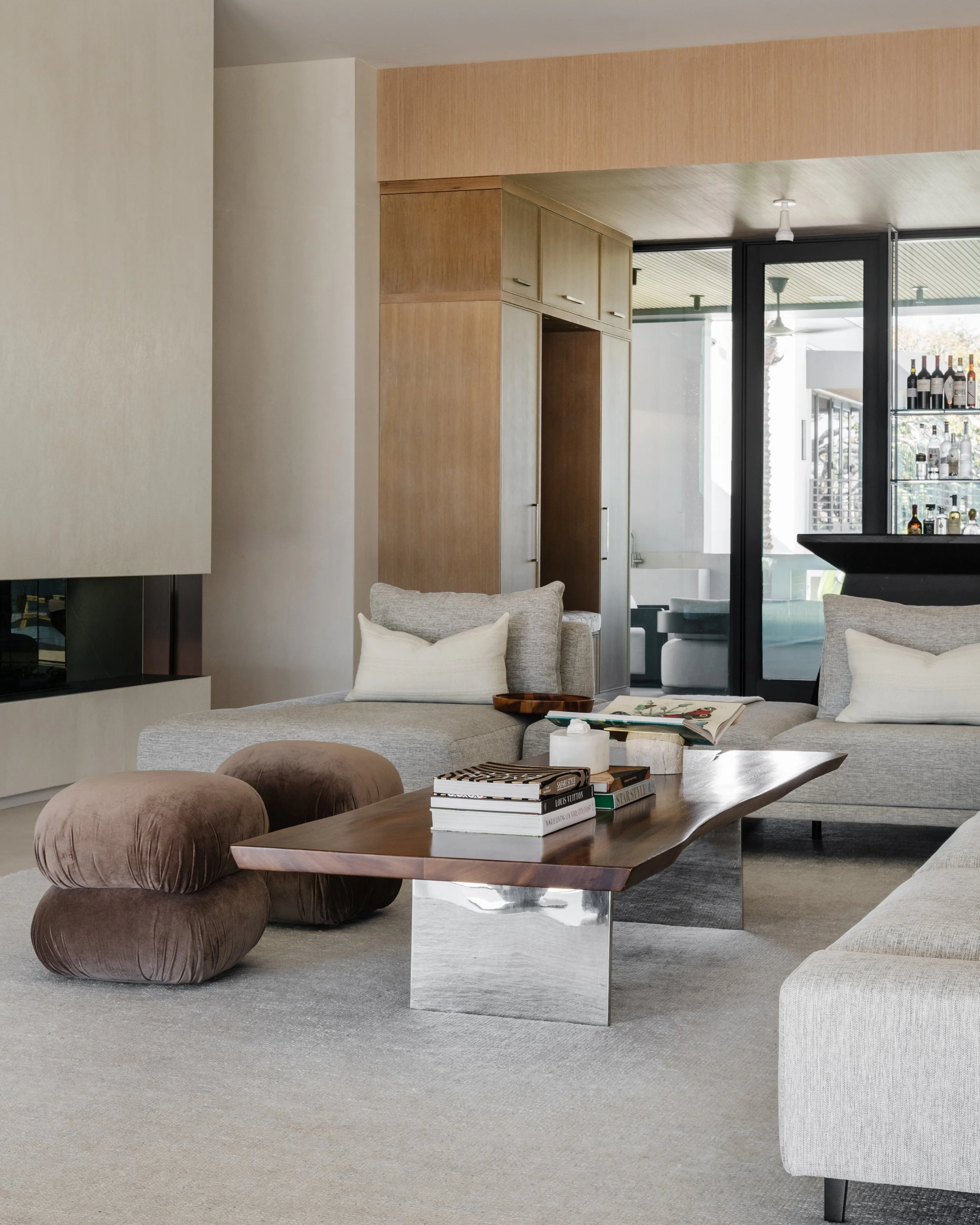A modern living room with beige and light gray furniture, a wooden coffee table, brown tufted ottomans, and shelves with wine bottles in the background.