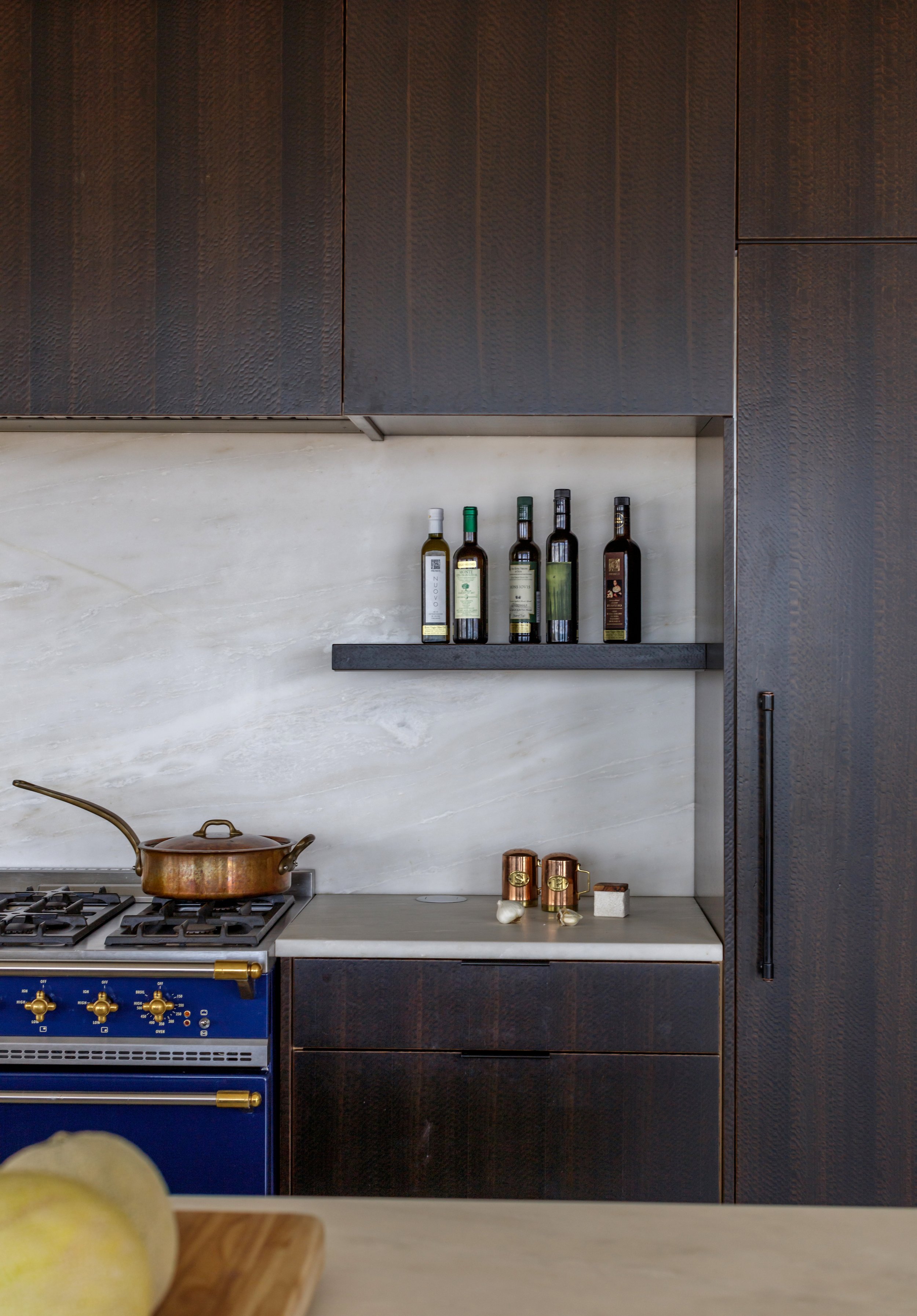 Modern kitchen with dark wooden cabinets, white marble backsplash, and blue oven. A copper pot is on the stovetop, and bottles of oil are on a shelf above the countertop. Copper mugs and garlic are on the counter.