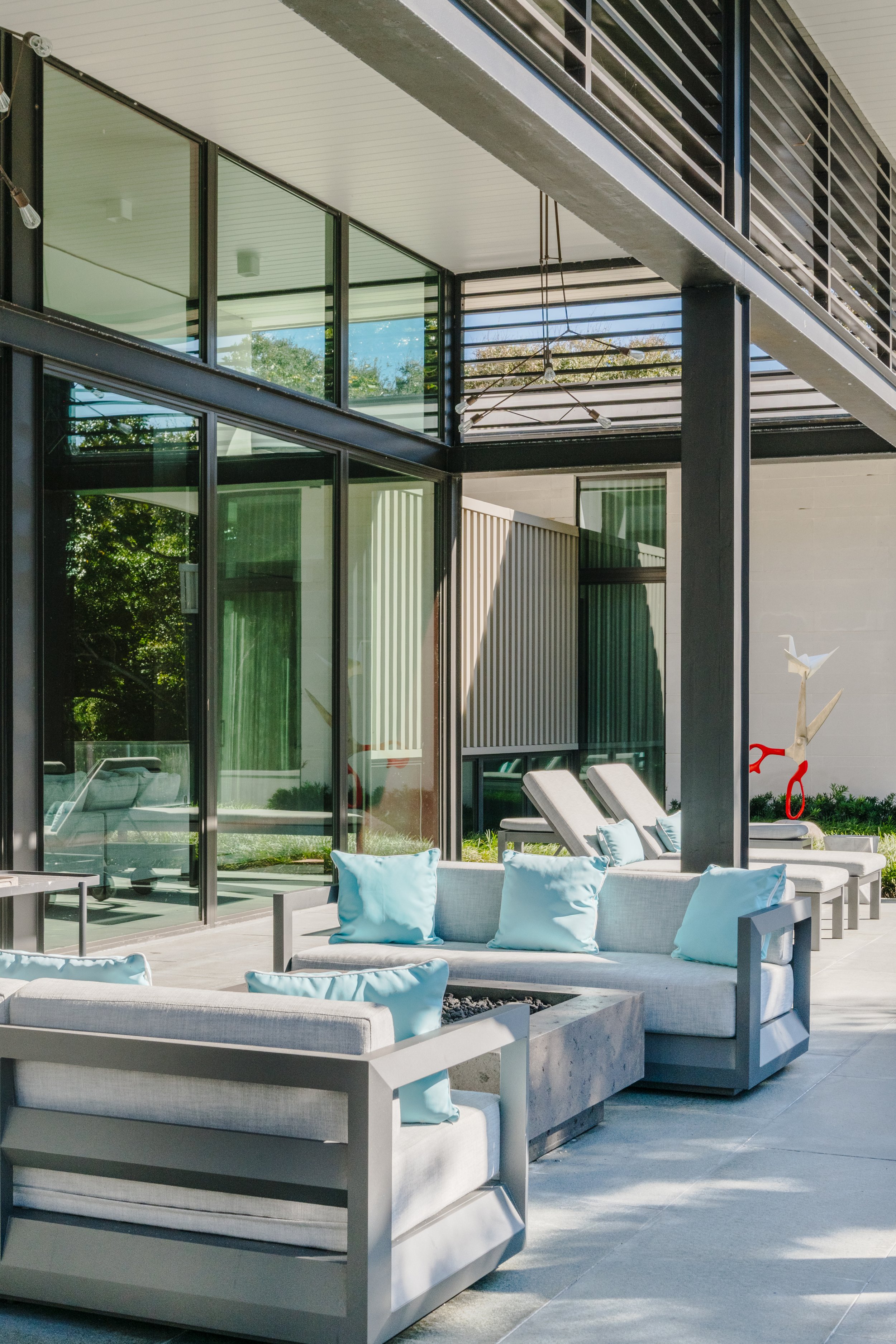 Modern outdoor patio with white cushioned seating, blue pillows, sun loungers, and a sculpture of scissors with red handles, adjacent to a contemporary building with large glass windows and metal railings.