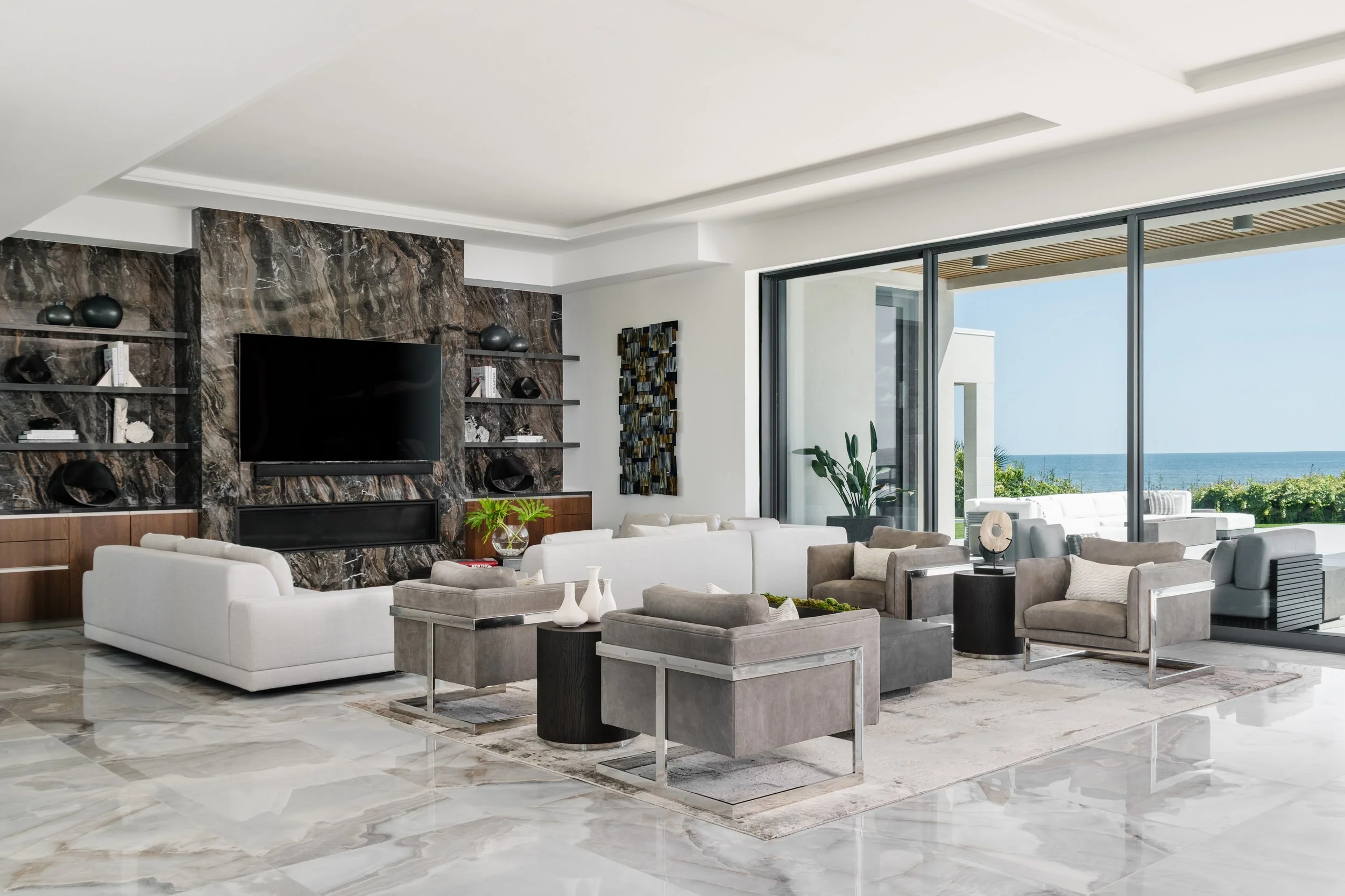 A modern living room with white and gray furniture, a marble fireplace surround, open shelves, and large windows showing an ocean view.