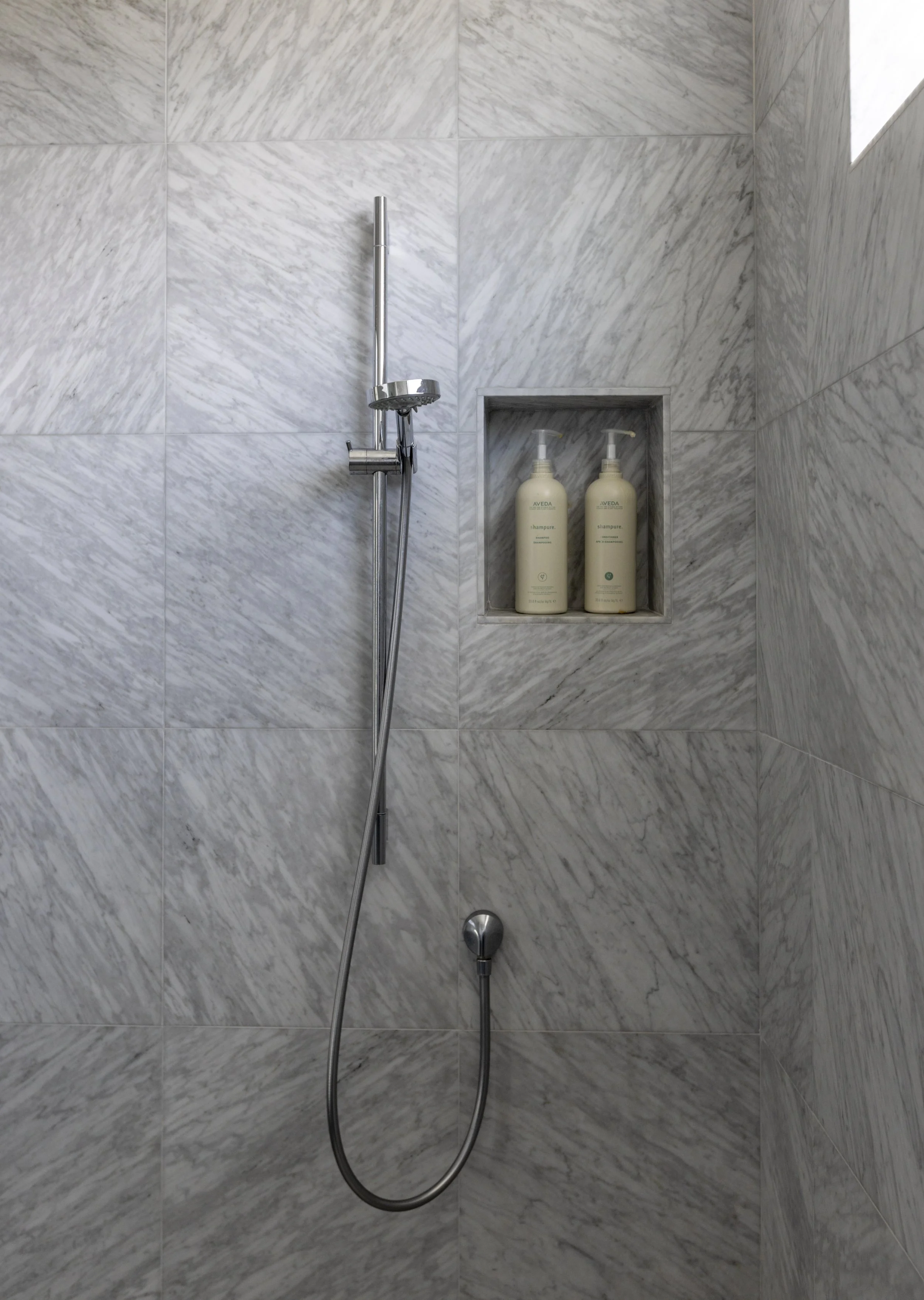 A shower with grey marble walls, a handheld showerhead, and a built-in shelf holding two bottles of shampoo and conditioner.