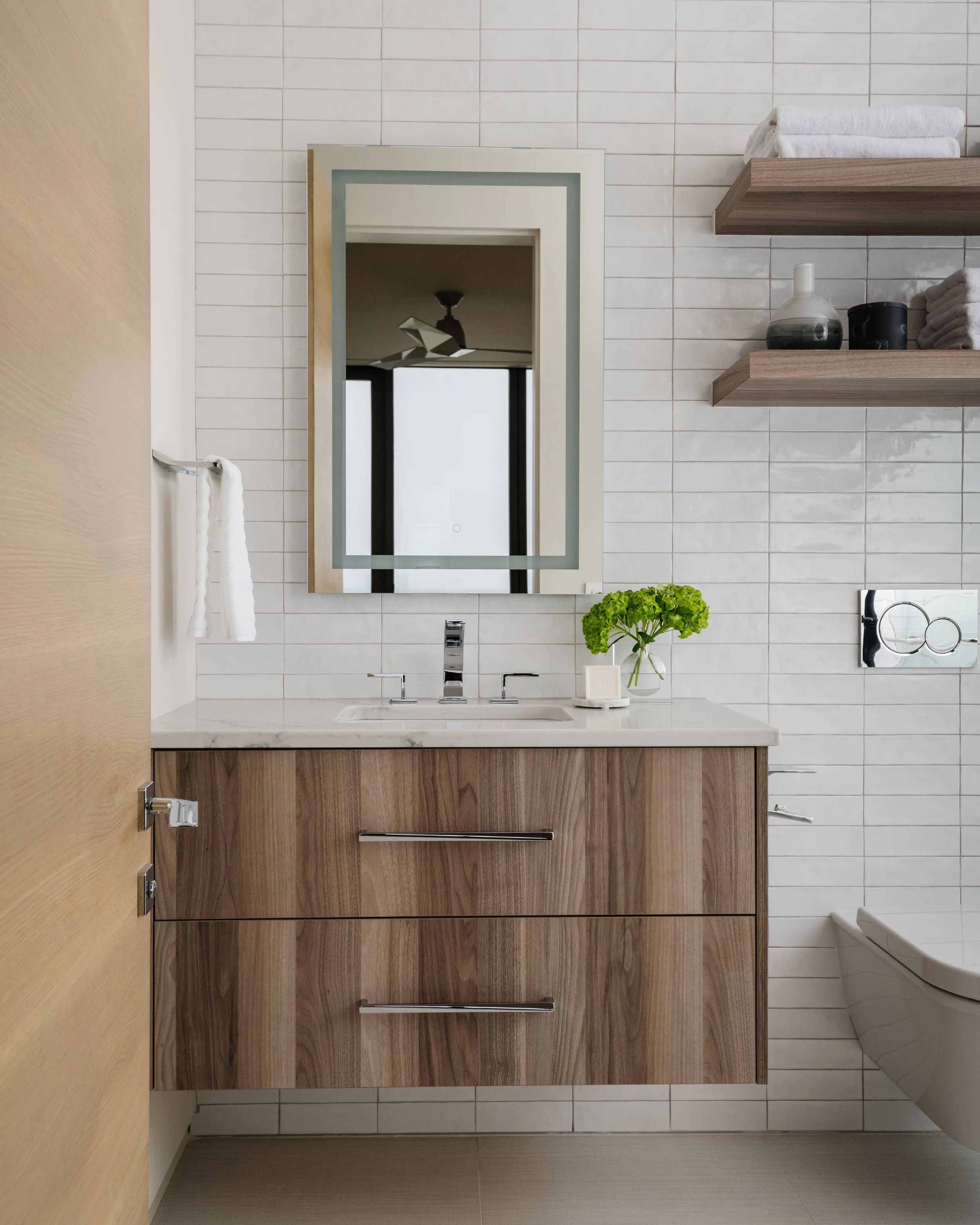 Modern bathroom with white tiled walls, wooden vanity with drawers, a large mirror, a glass vase with green flowers, and two floating shelves holding folded towels and decorative items.
