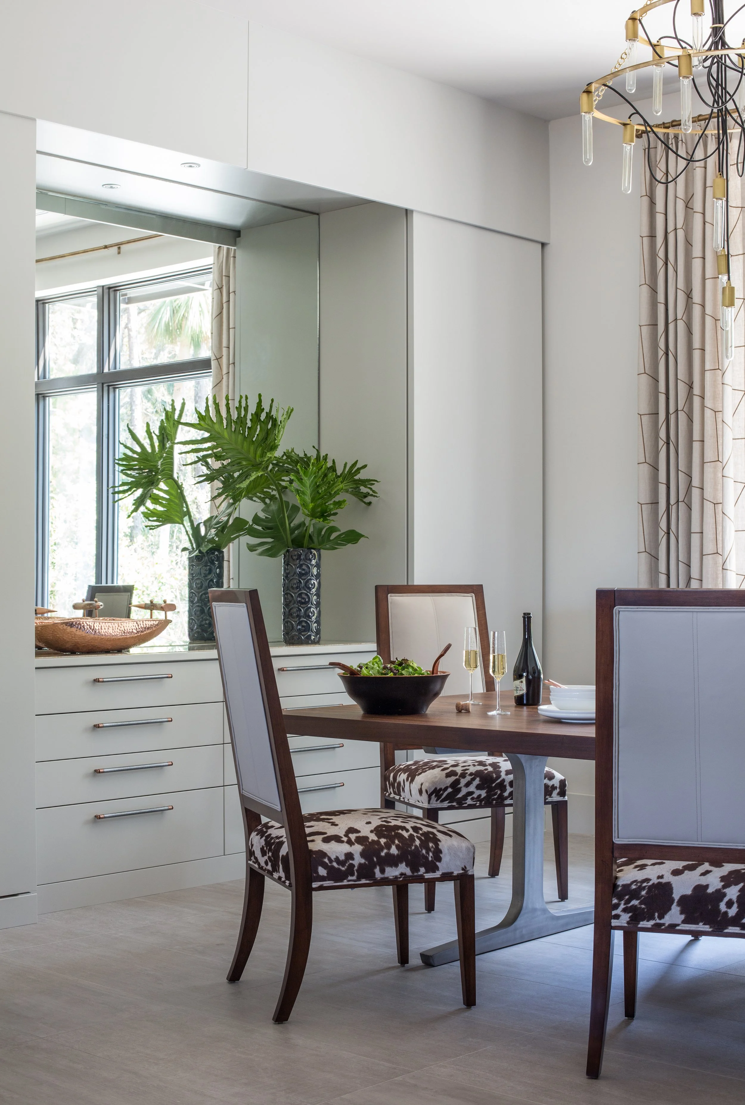 A dining room with a wooden table set with wine glasses, a bottle of wine, and a salad bowl. There are chairs with animal print upholstery and white backs. A large window with curtains and a potted plant are visible, along with a modern chandelier.
