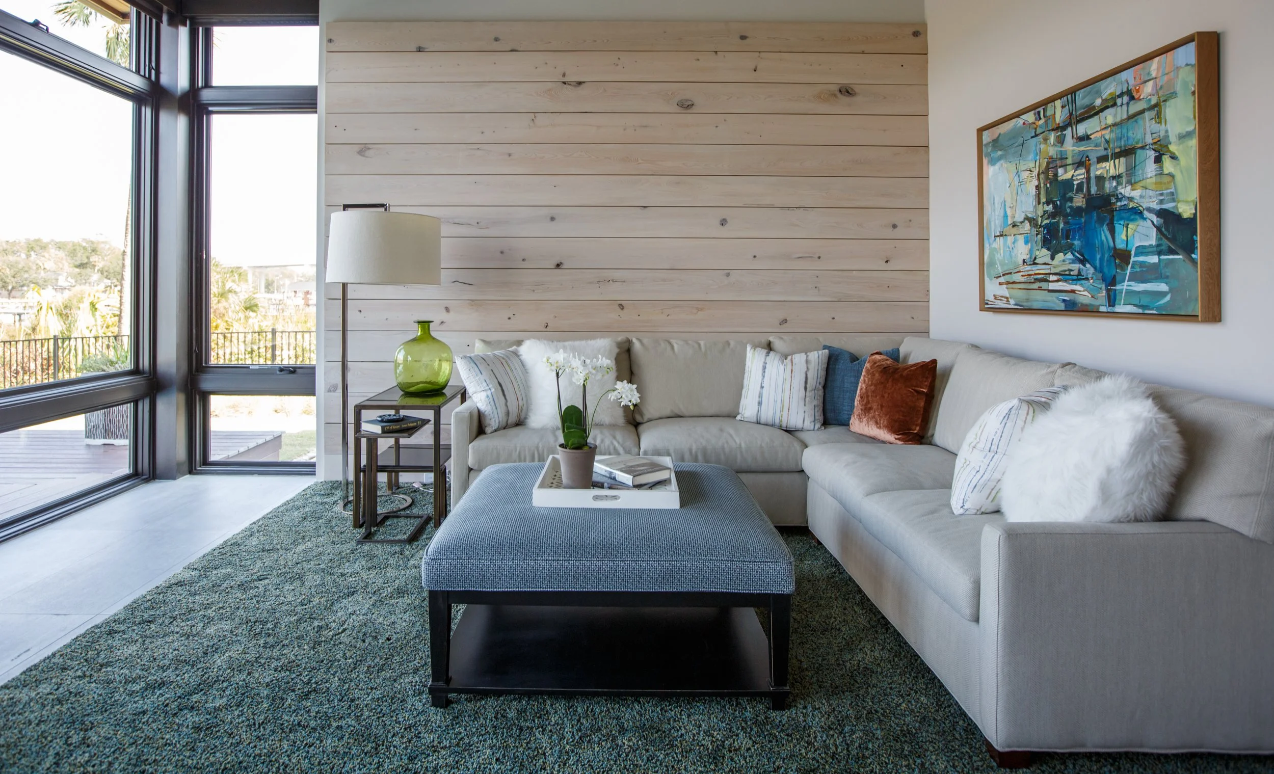 Living room with large windows, a sectional sofa with pillows, a coffee table with a plant and books, a green vase, a floor lamp, a textured green rug, a wood-paneled wall, and a colorful abstract painting on the wall.