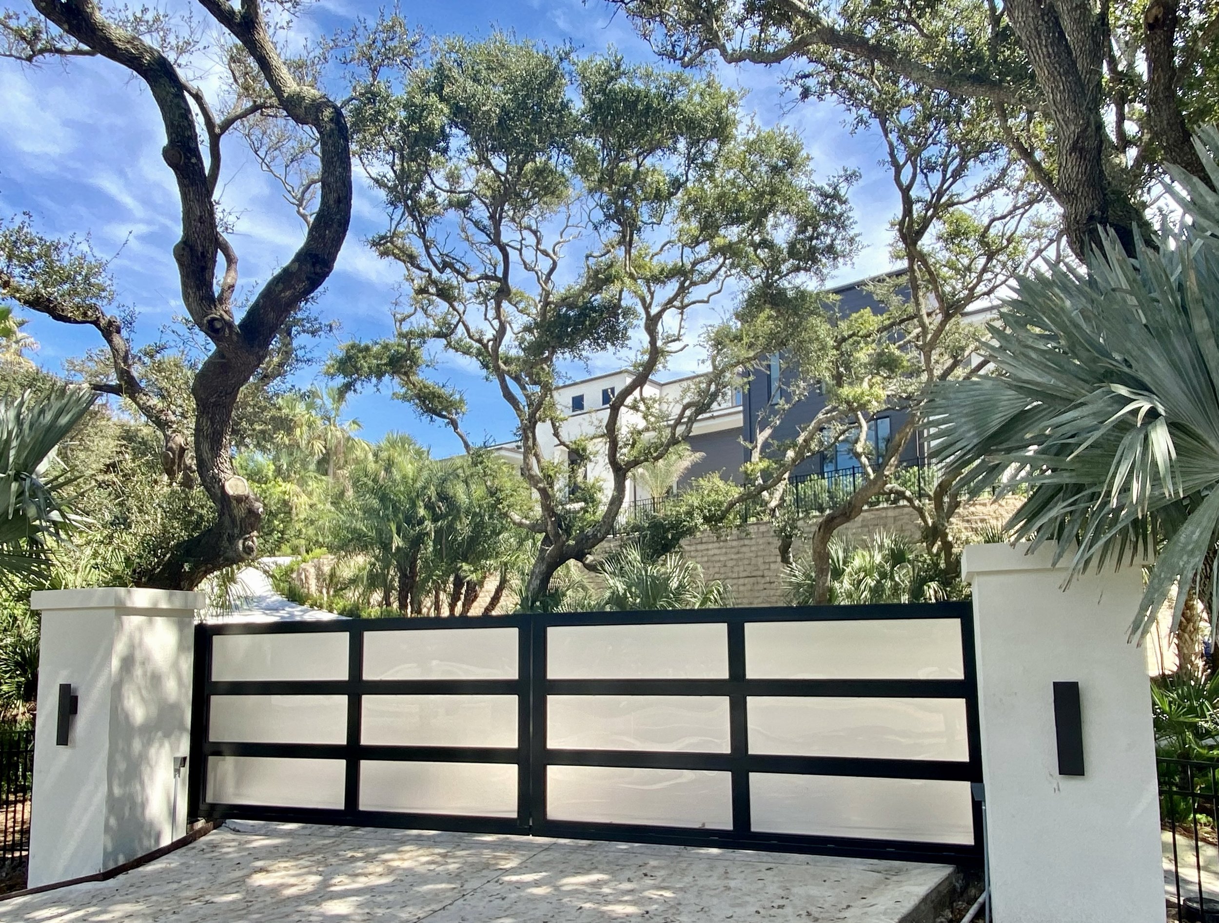 A black and white modern gate with grid pattern in front of a garden with trees and a building in the background under a blue sky with clouds.