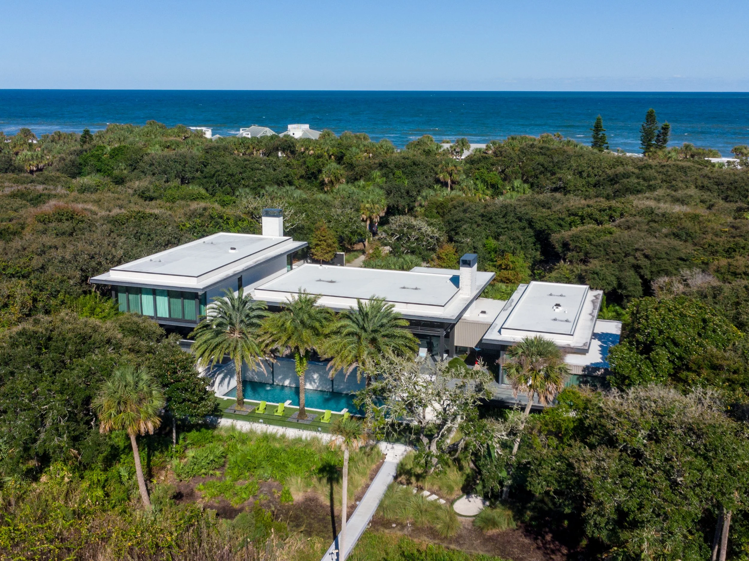 Aerial view of a modern house with a flat roof, surrounded by lush green trees, with the ocean in the background under a clear blue sky.