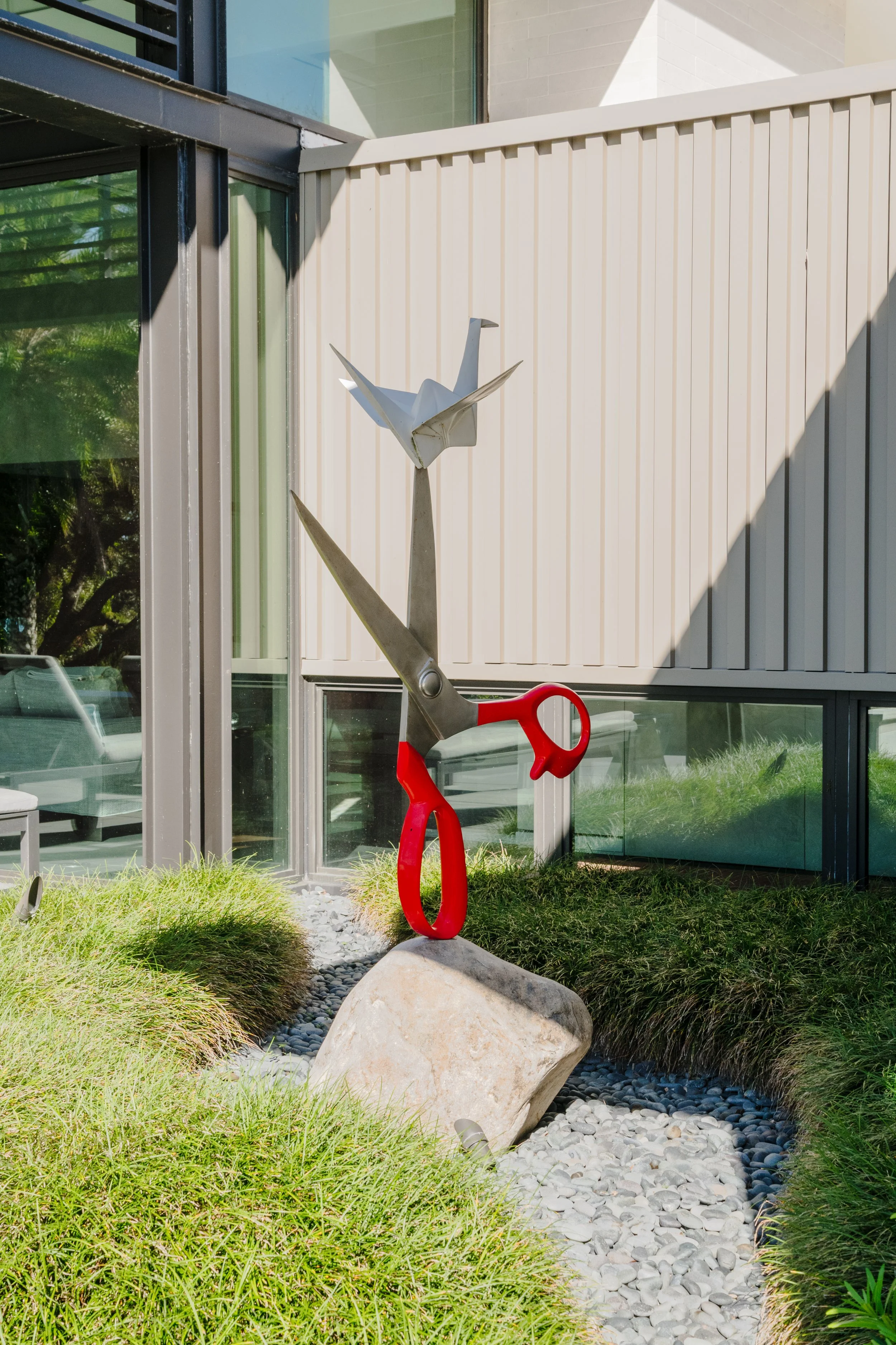 An outdoor modern art sculpture of a bird with outstretched wings perched on a red scissors-shaped stand, located in a landscaped area with green grass, rocks, and a concrete boulder, next to a building with large glass windows and beige siding.
