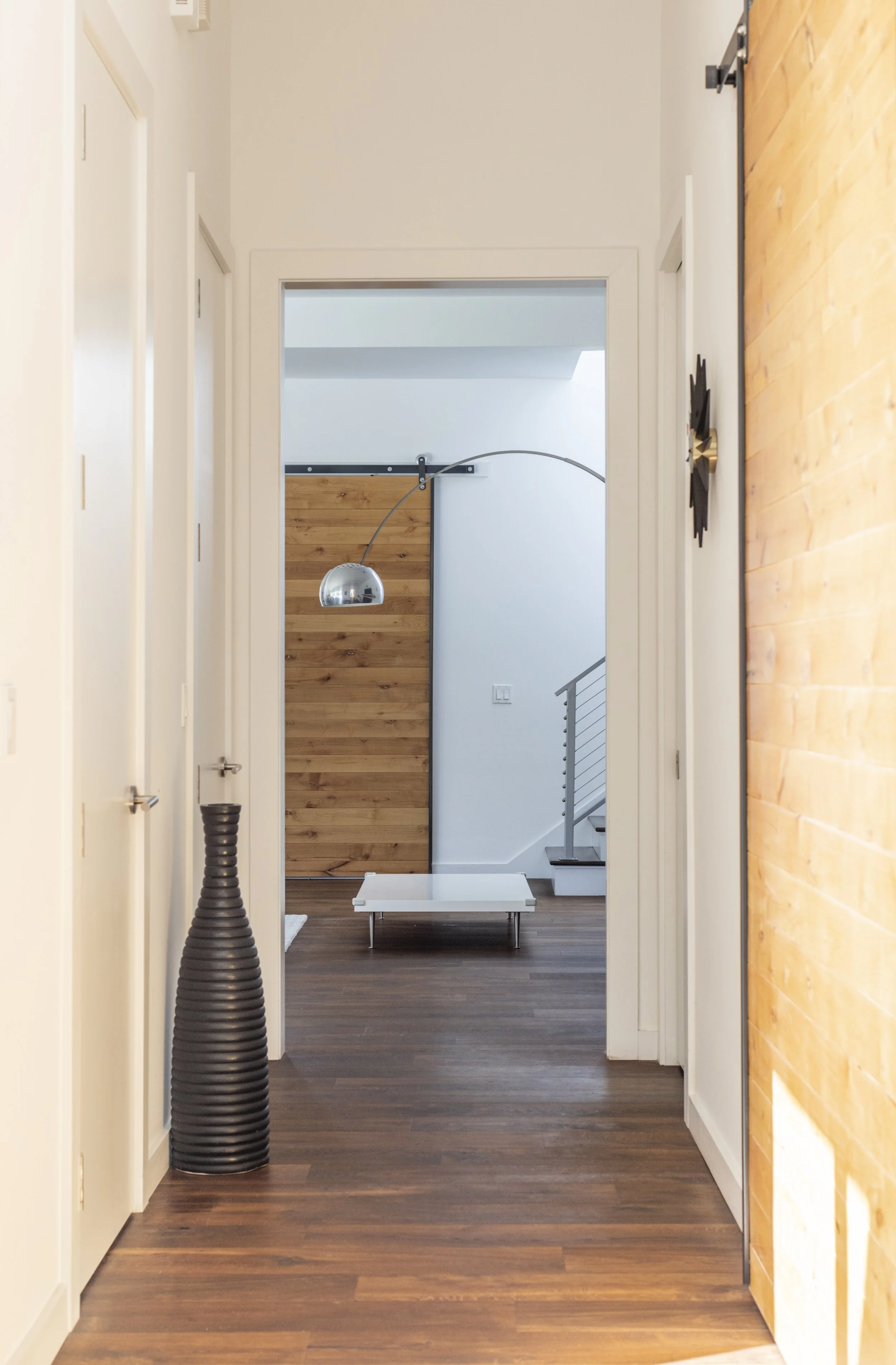 View of a hallway with wooden floors and white walls, a large black vase on the left, a white bench at the end, and a wooden sliding barn door on the right.